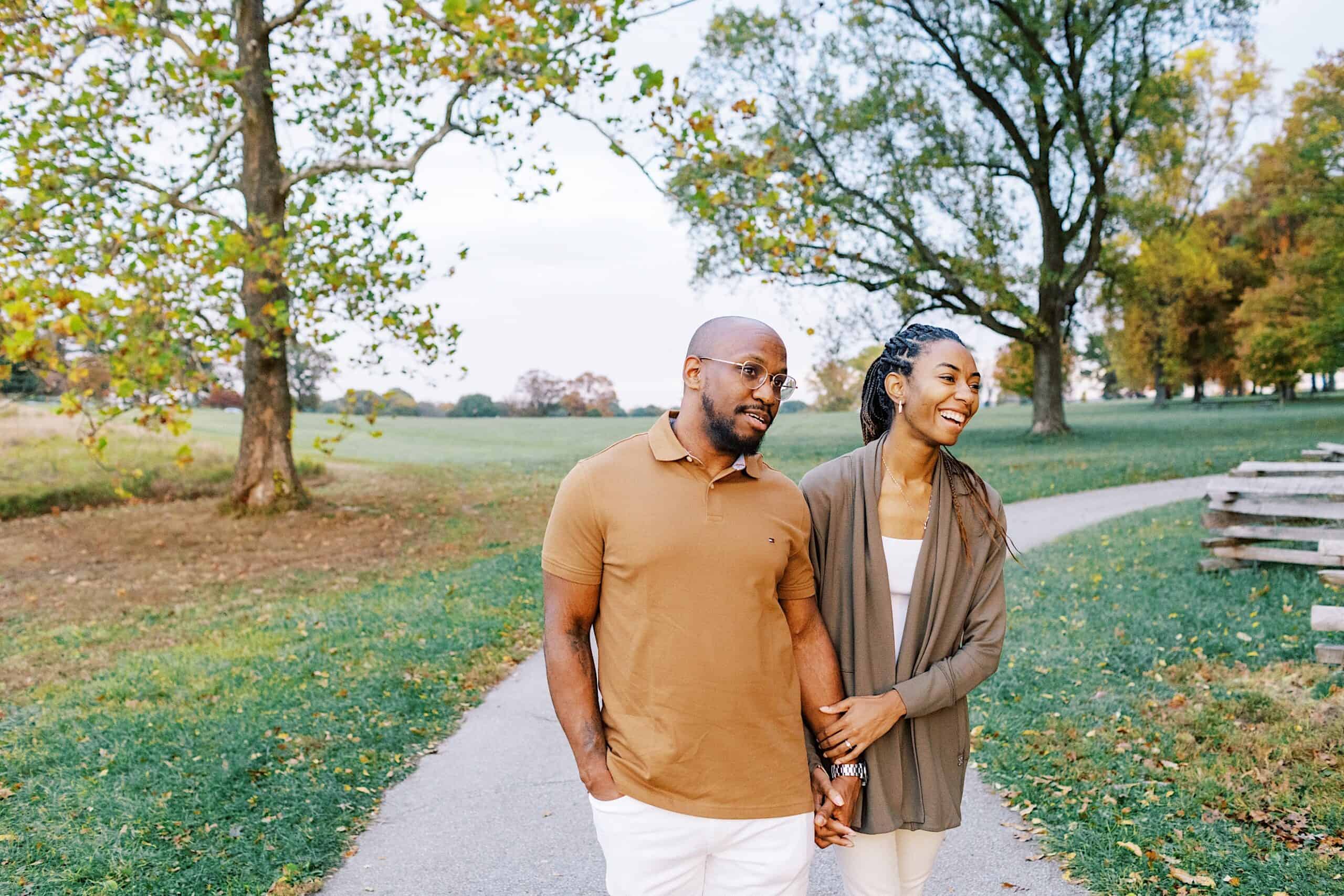 A man and woman walk hand in hand along a paved path in a park surrounded by green grass and trees.