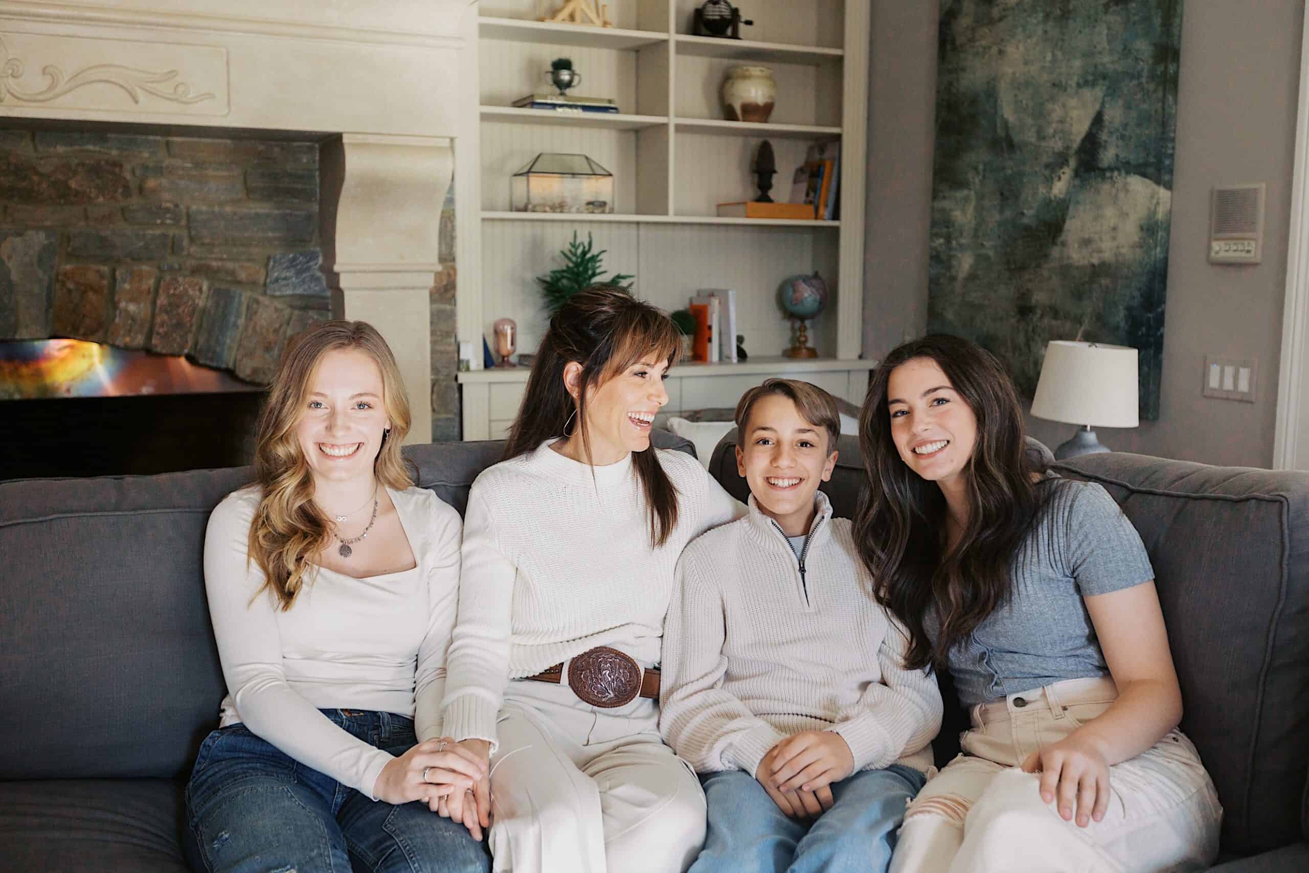 Four people sit closely together on a gray couch, smiling at the camera; there are bookshelves and a fireplace in the background.