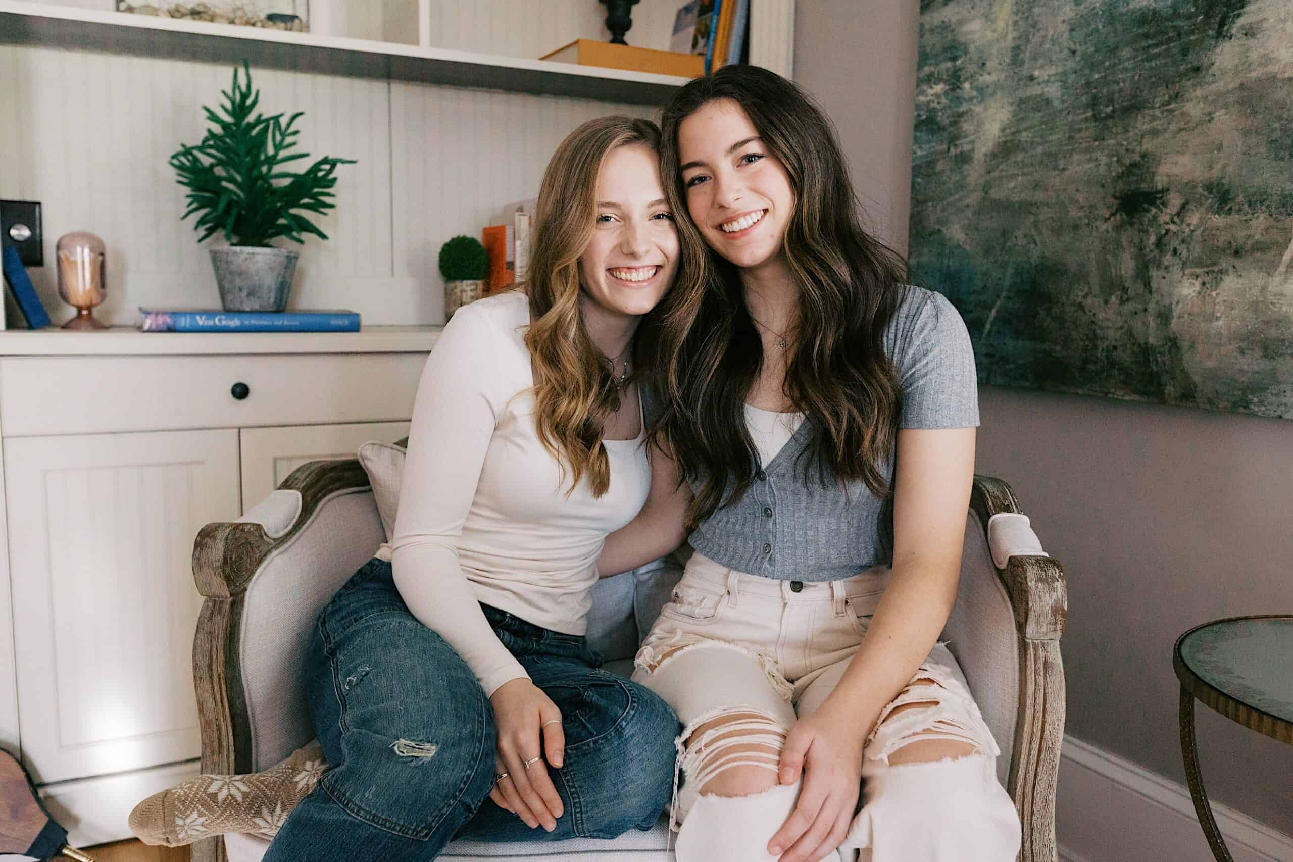 Two young women sit close together on a chair, smiling at the camera in a cozy living room setting with shelves, books, and decor in the background.