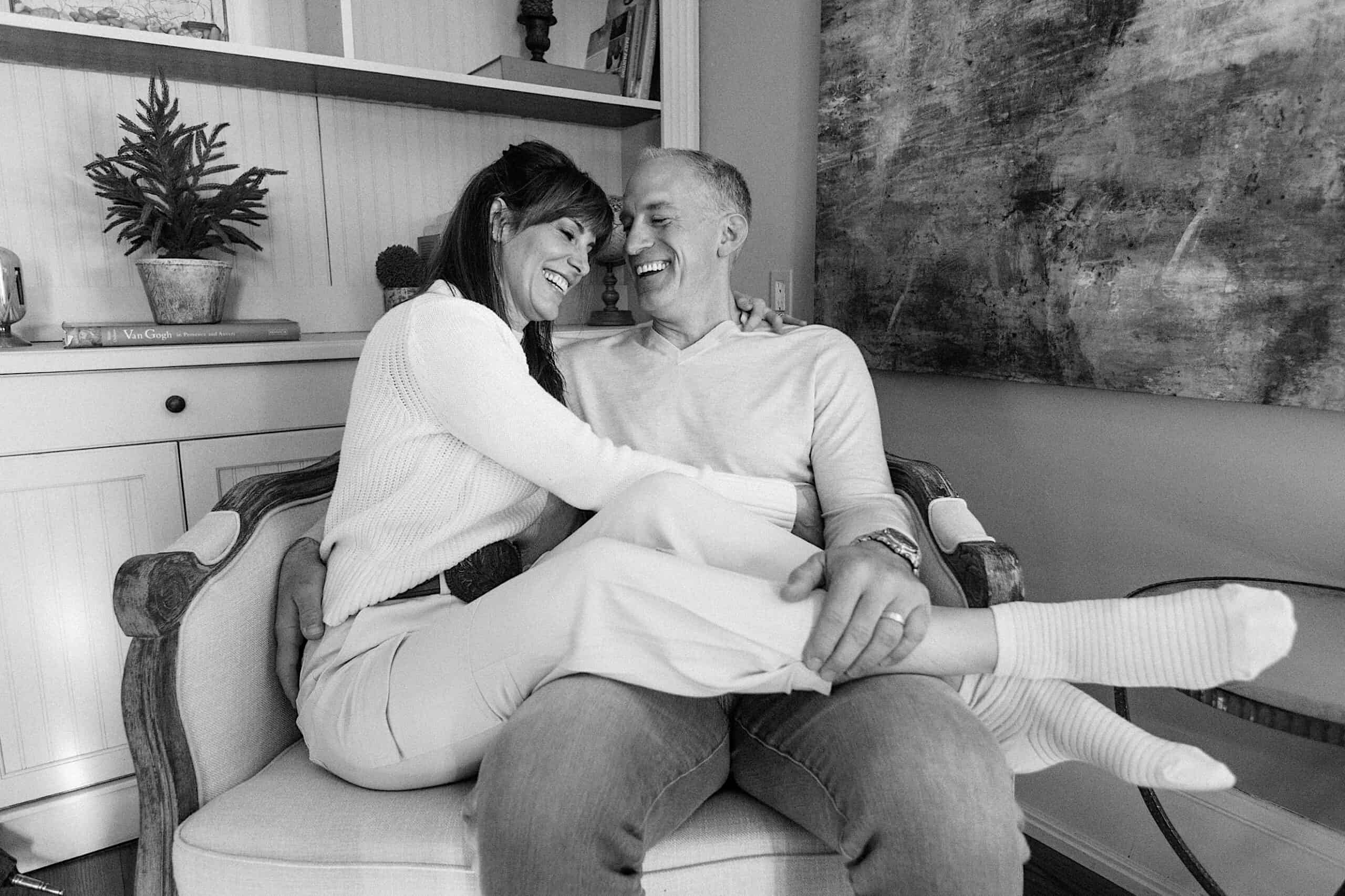 A man and woman sit closely together in an armchair, smiling and laughing in a cozy living room.