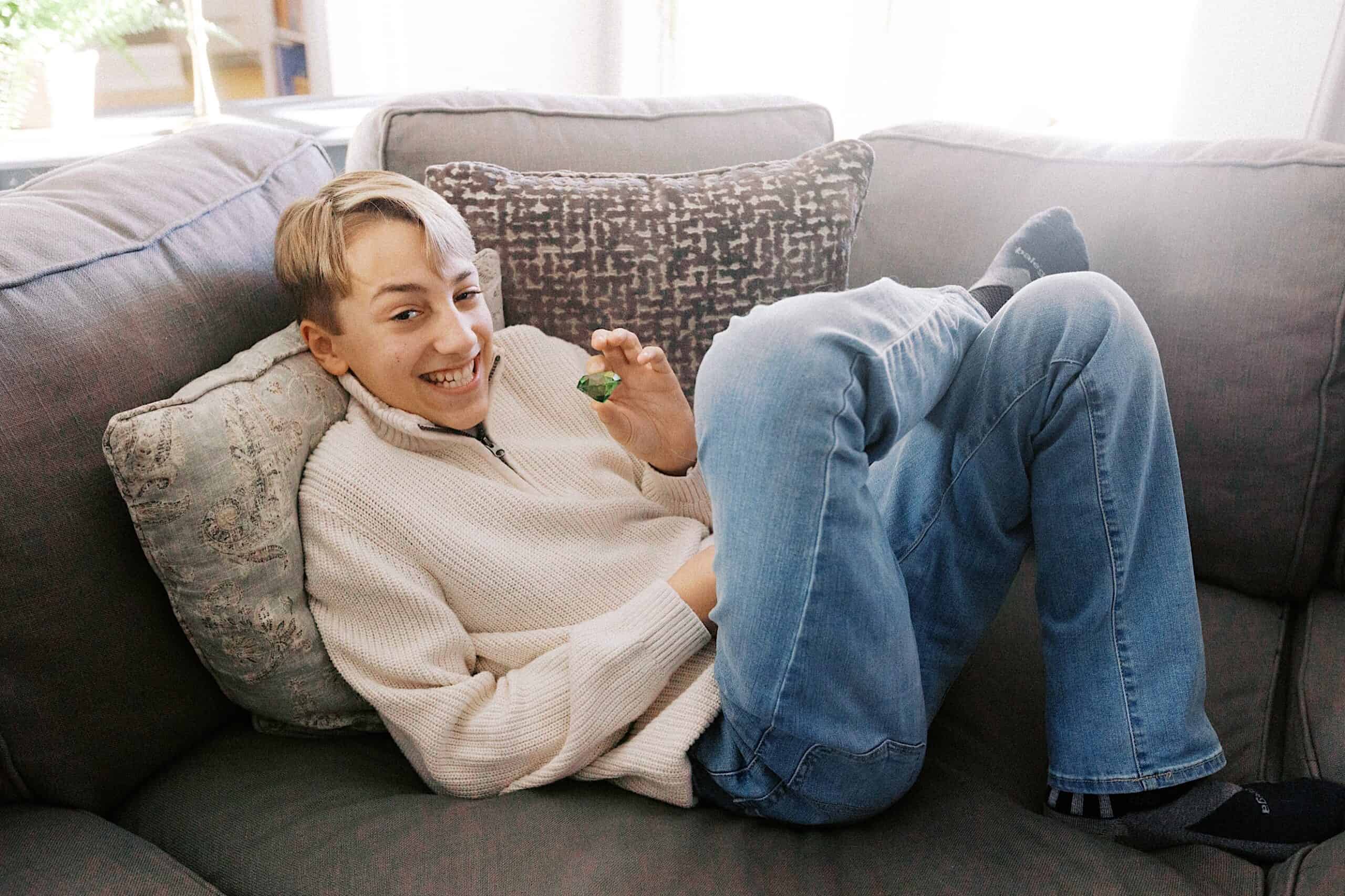 A young person in a cream sweater and blue jeans sits on a gray couch, smiling and holding a small green object in their hand.