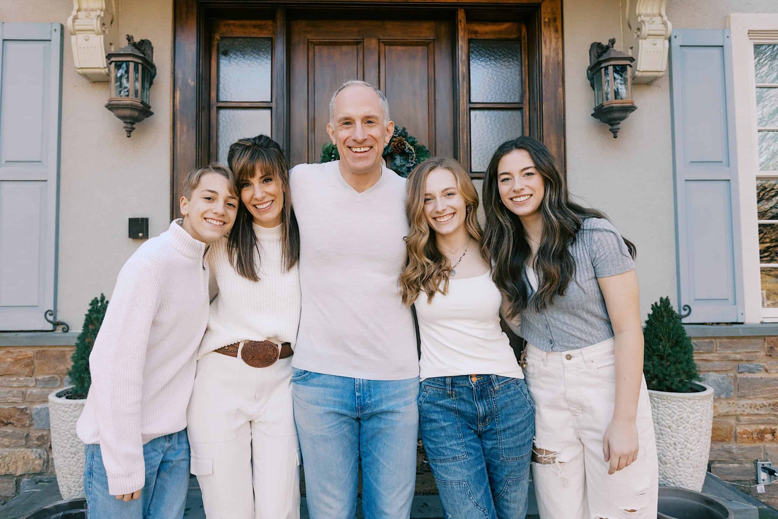 Five people stand closely together smiling in front of a house with wooden doors and potted plants on either side.