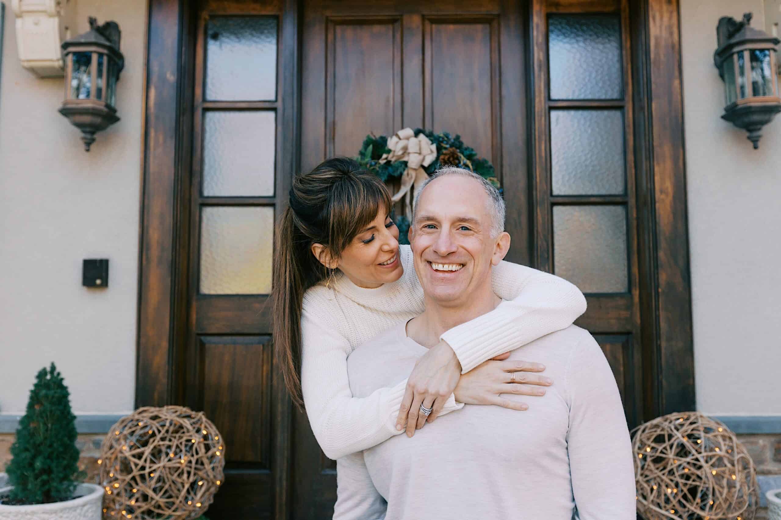 A woman embraces a smiling man from behind in front of a wooden door decorated with a wreath. Decorative lights and small plants are visible on either side of the doorway.