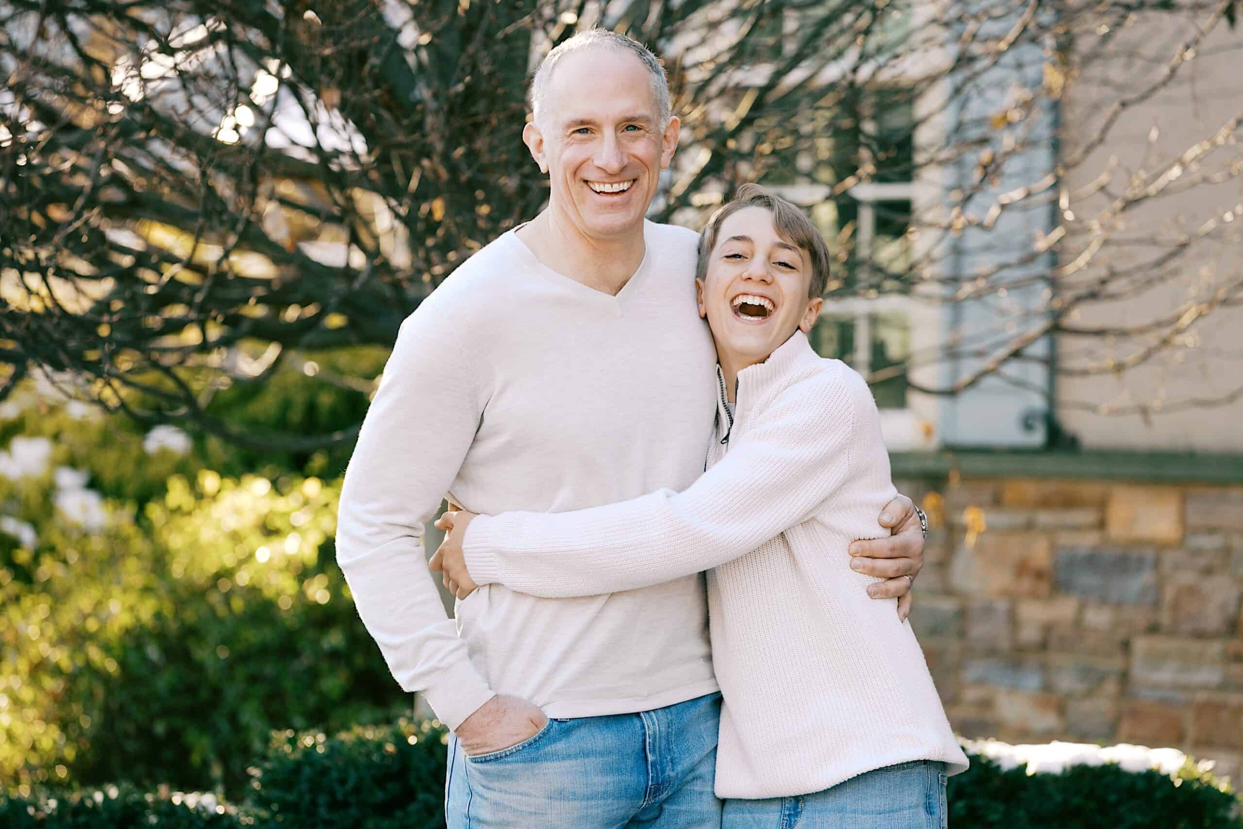 Two people stand outdoors in front of a tree and building, smiling and hugging each other while wearing light-colored sweaters and jeans.