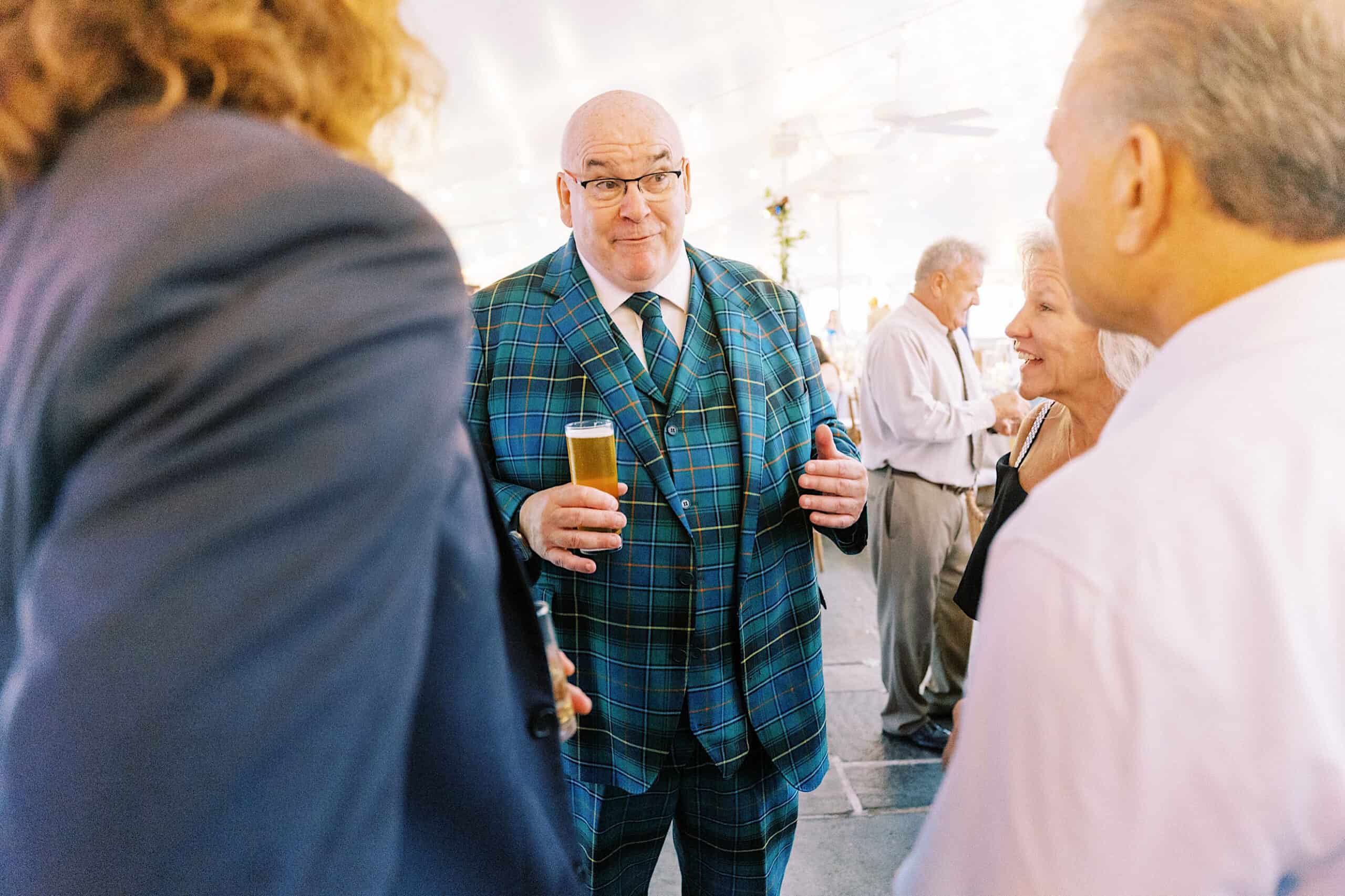 At a Summer Wedding at Appleford Estate, a bald man in a blue plaid suit holds a glass of beer and chats with three people at a lively social gathering.