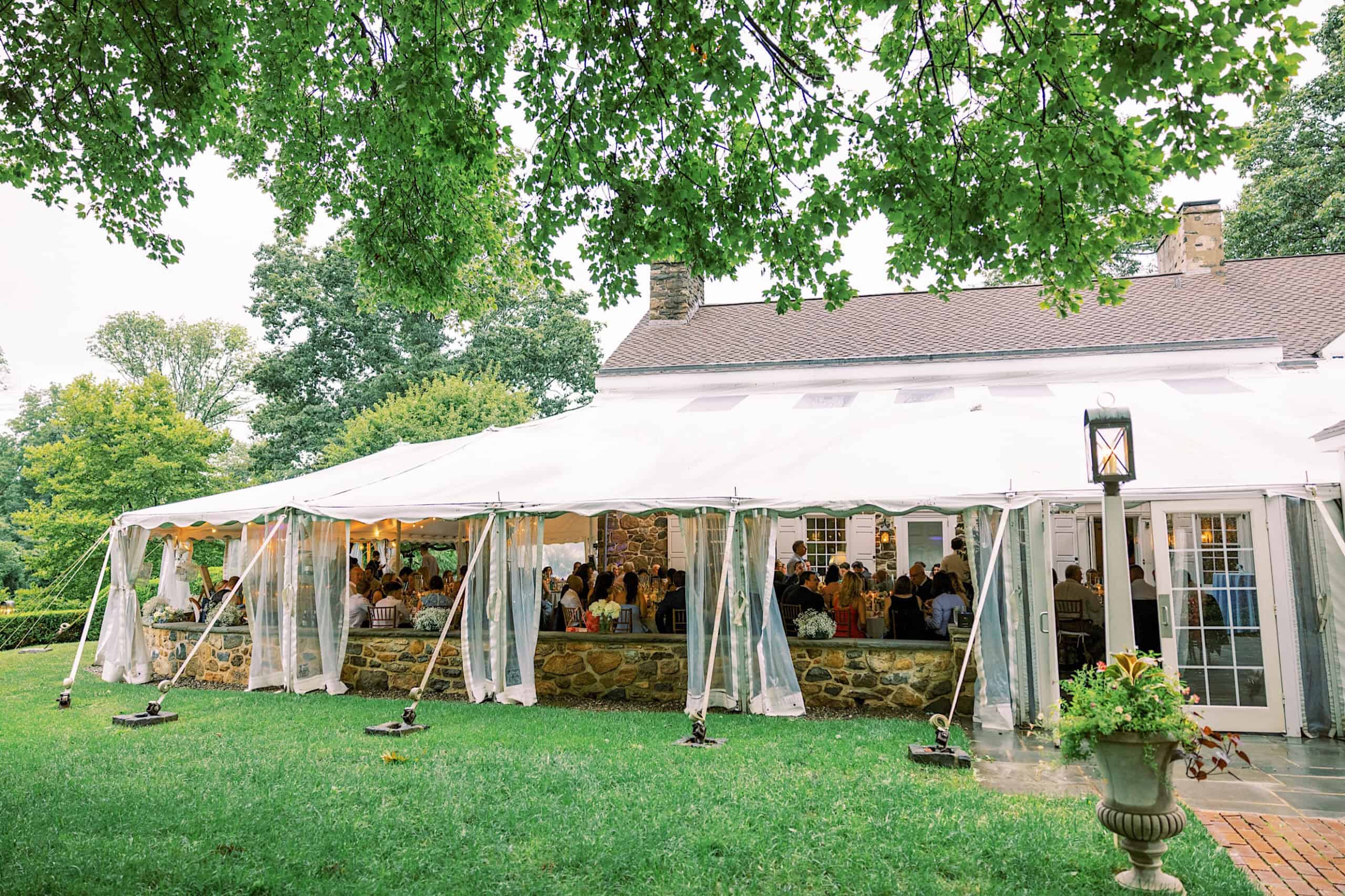 A white event tent set up beside a stone house hosts guests seated at tables; green lawn and trees surround the area, creating a picturesque Summer Wedding at Appleford Estate.
