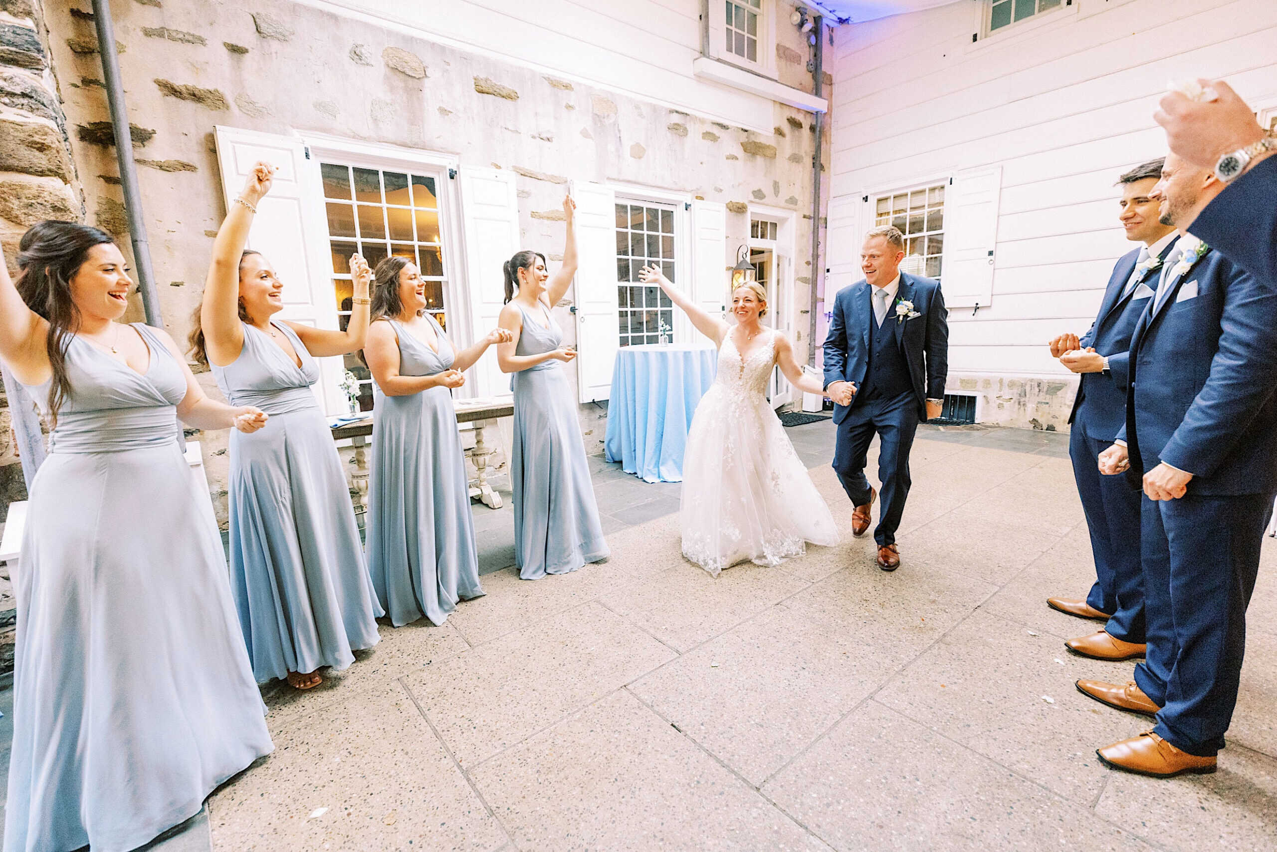 At a Summer Wedding at Appleford Estate, a bride and groom enter the courtyard holding hands, greeted by cheering bridesmaids in light blue dresses and groomsmen in blue suits.
