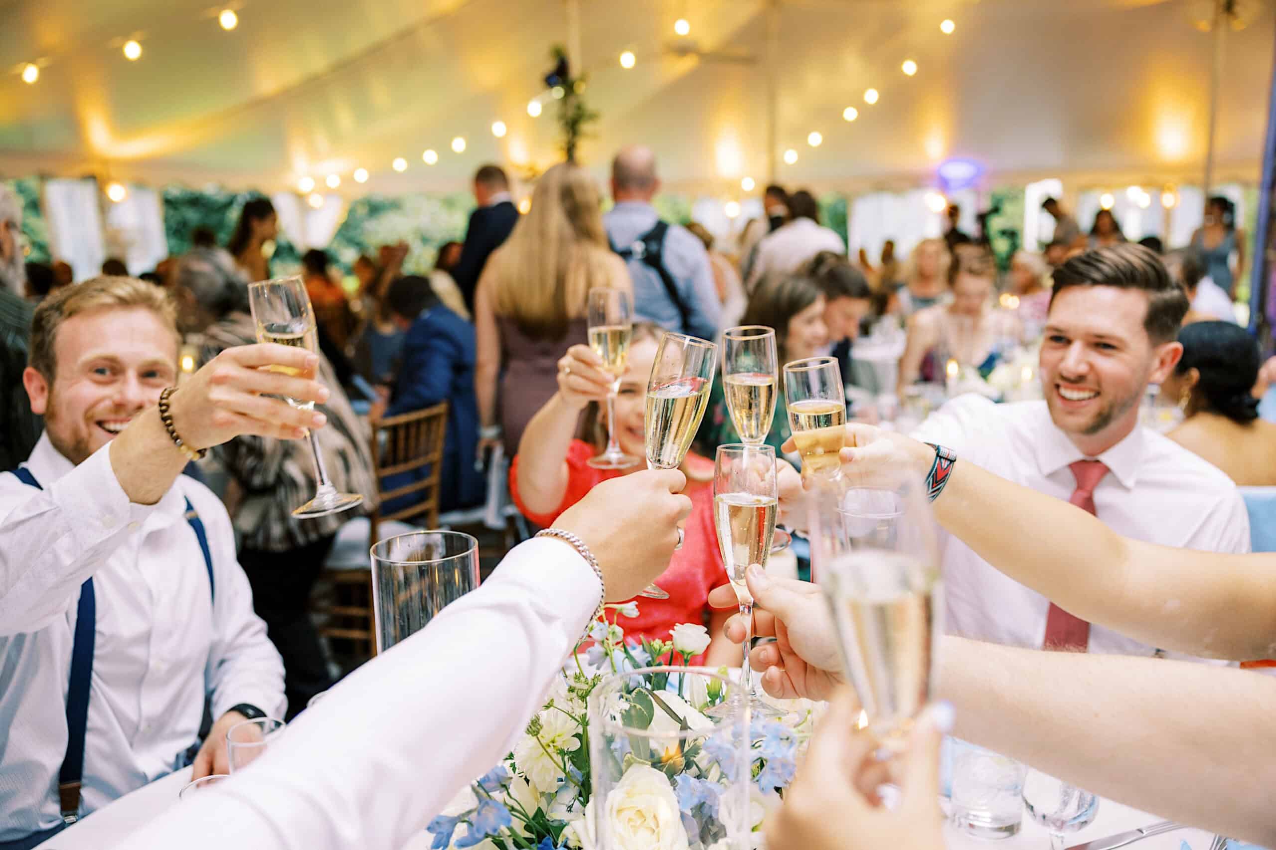 A group of people raise champagne glasses in a toast at a Summer Wedding at Appleford Estate, celebrating under a decorated tent with glowing string lights.