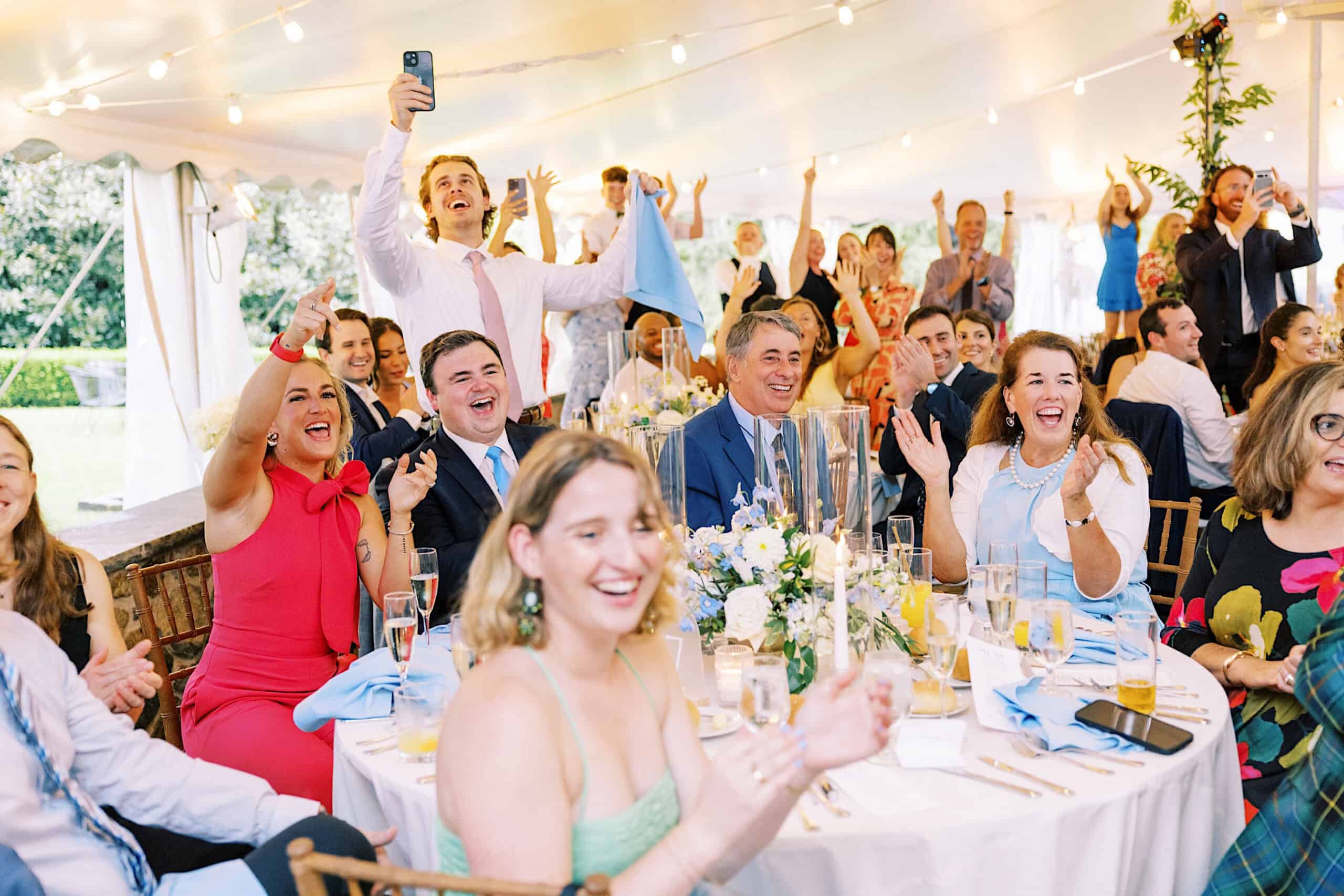 At a Summer Wedding at Appleford Estate, well-dressed guests sit and stand around banquet tables in a decorated tent, cheering and smiling, with raised hands and phones capturing the joyful moment.