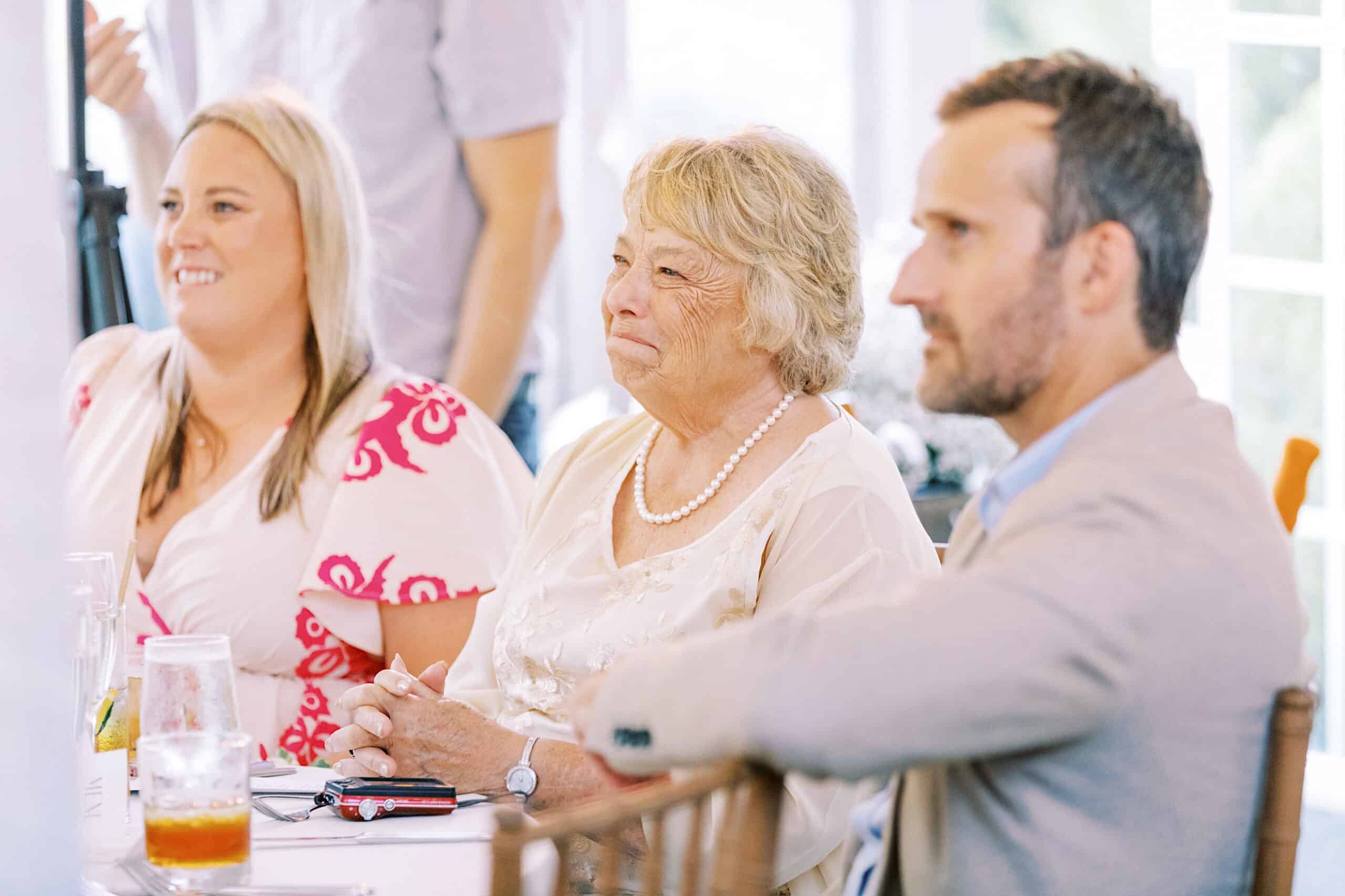 Three people sit at a table in a well-lit indoor setting, attentively watching something off-camera during a Summer Wedding at Appleford Estate.