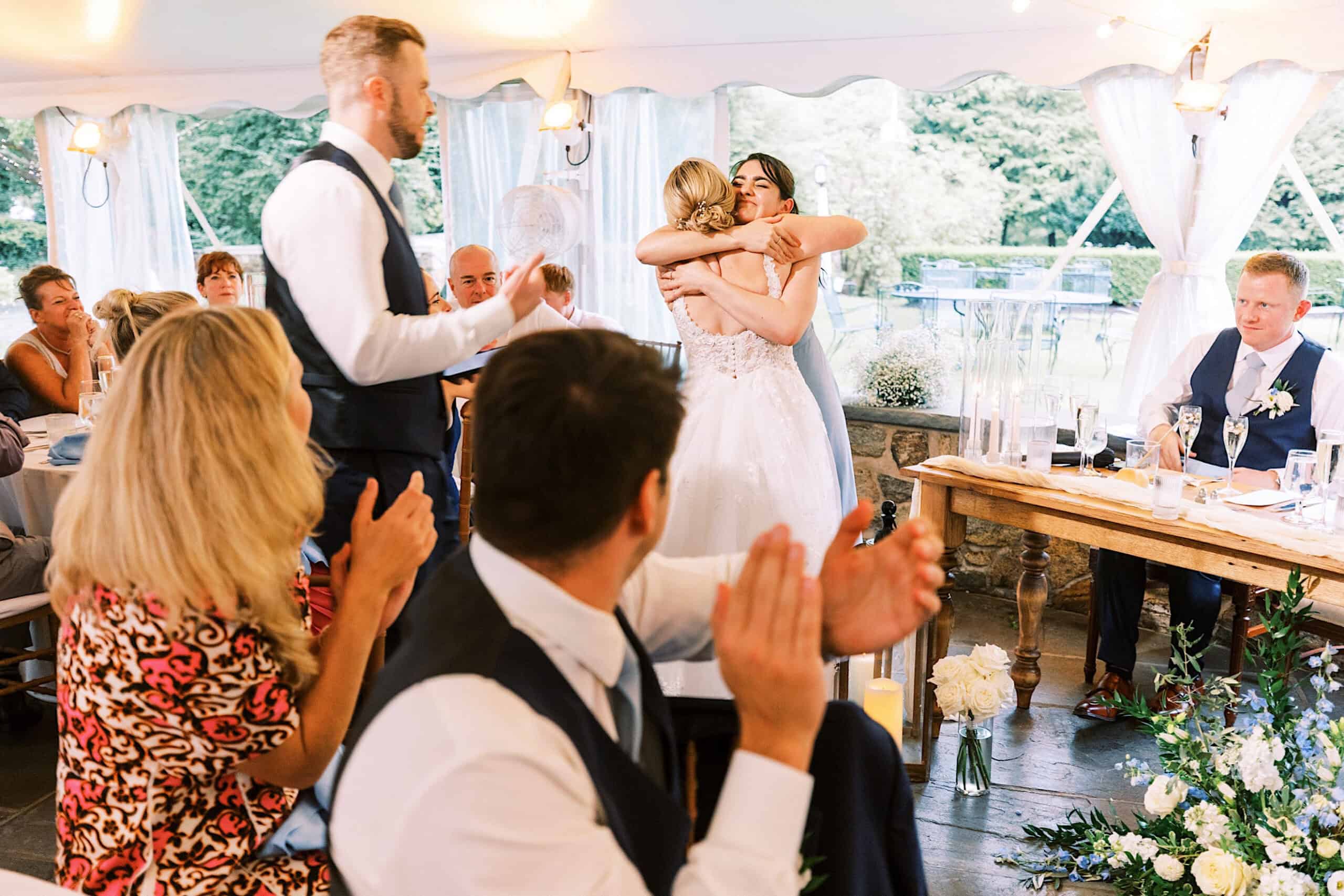 Two women in wedding attire hug while guests seated at tables watch and applaud inside a flower-filled, candlelit tented venue during a Summer Wedding at Appleford Estate.