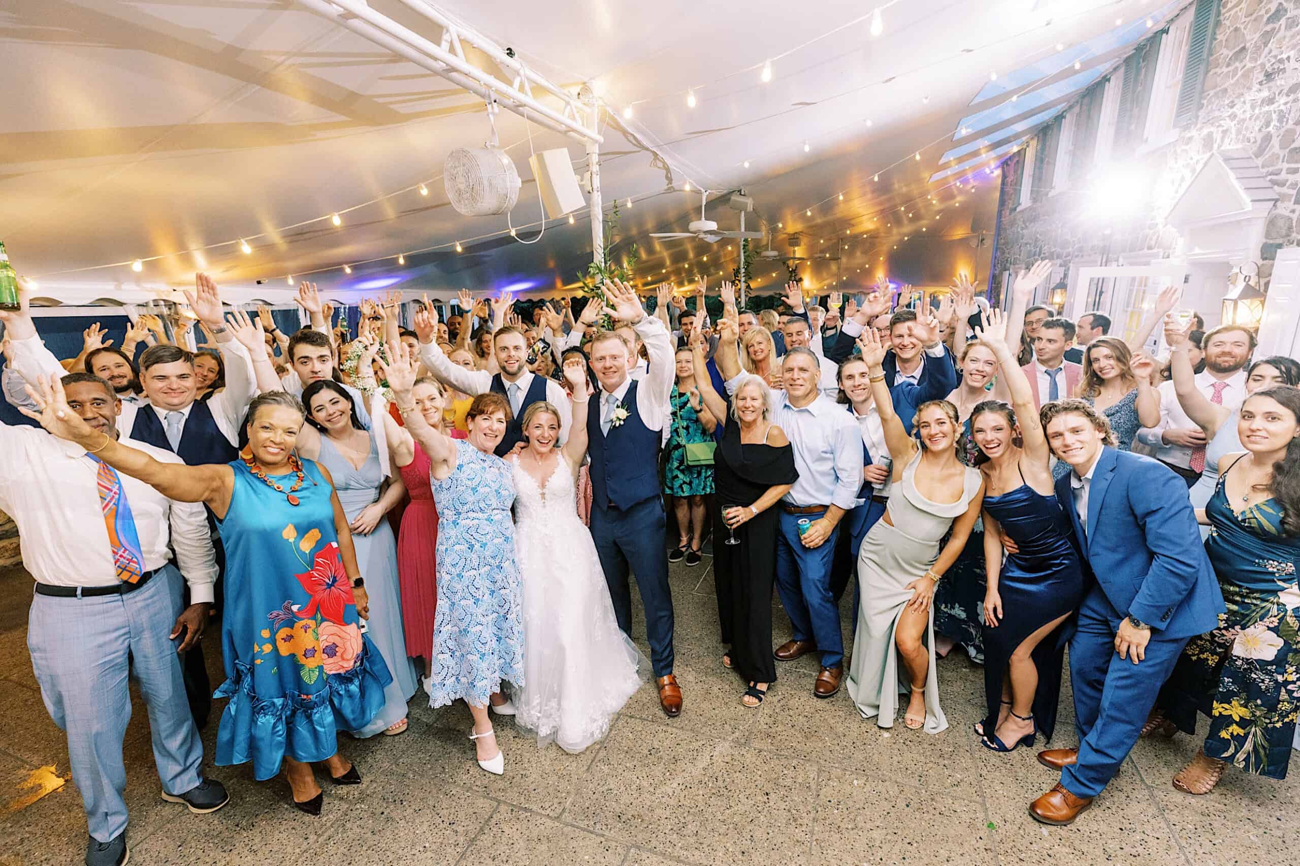 A large group of people, including a bride and groom, pose indoors at a joyful Summer Wedding at Appleford Estate reception with their hands raised, smiling at the camera.