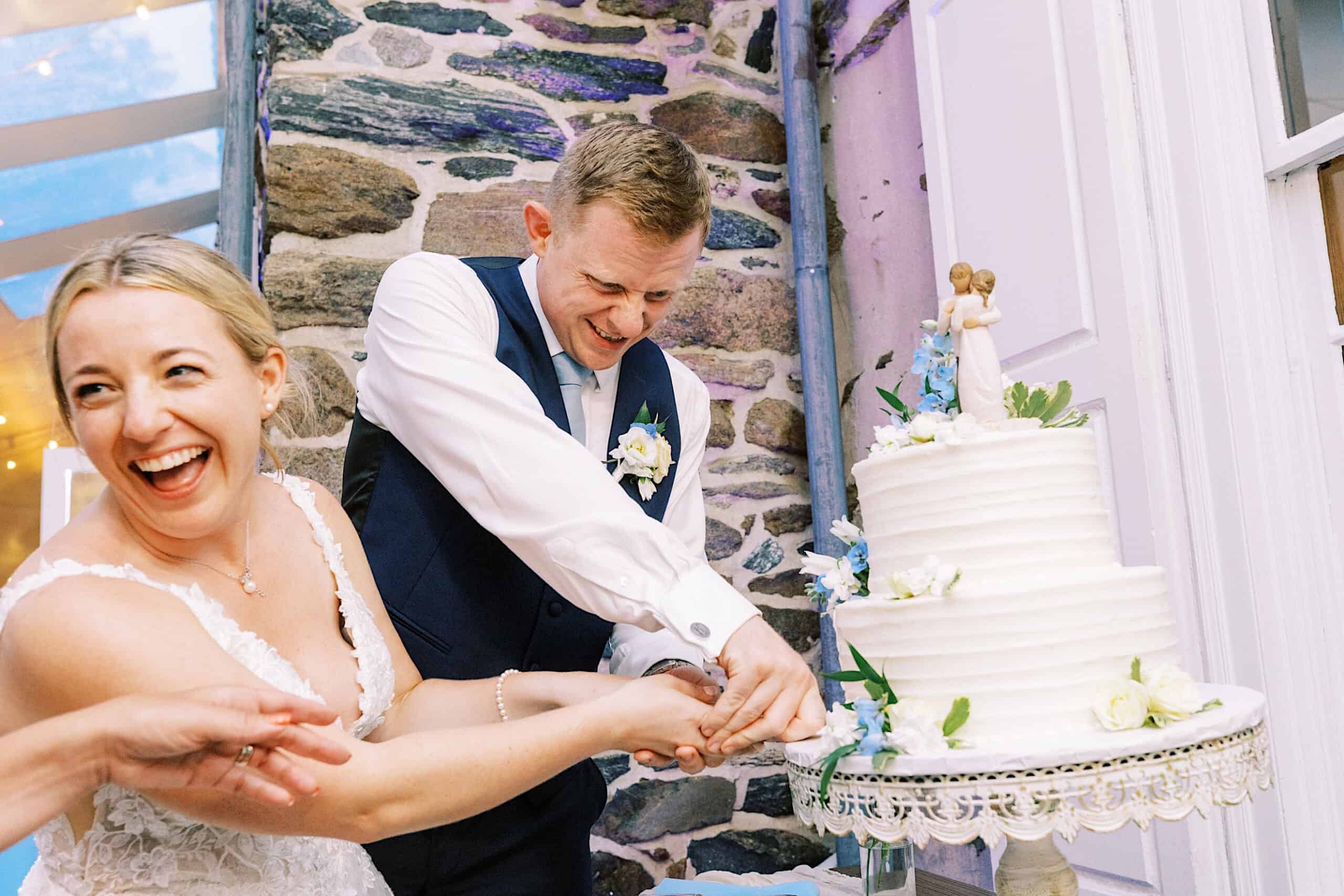 Bride and groom smiling and cutting a white tiered wedding cake together inside a stone-walled venue during their Summer Wedding at Appleford Estate.