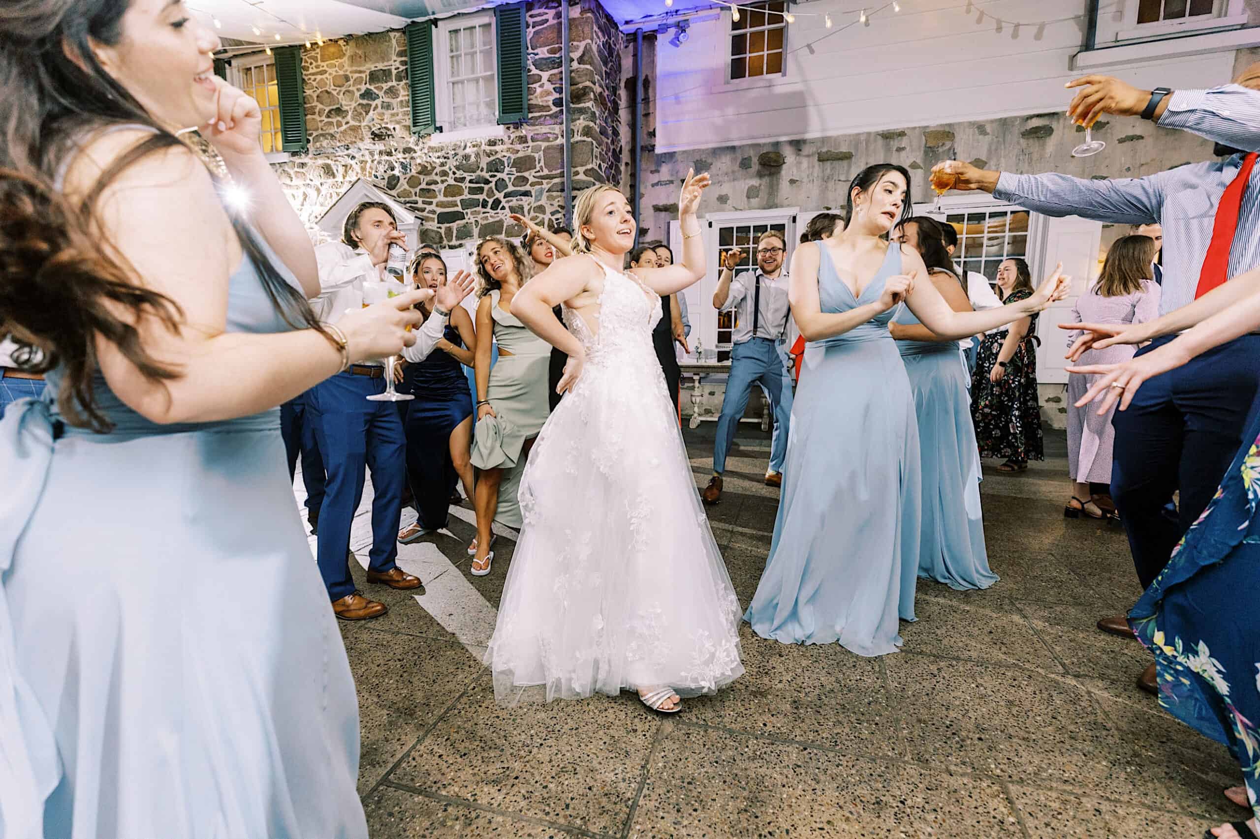 At a Summer Wedding at Appleford Estate, a bride in a white dress and guests in formal attire dance energetically at an indoor reception, surrounded by stone walls and twinkling string lights.