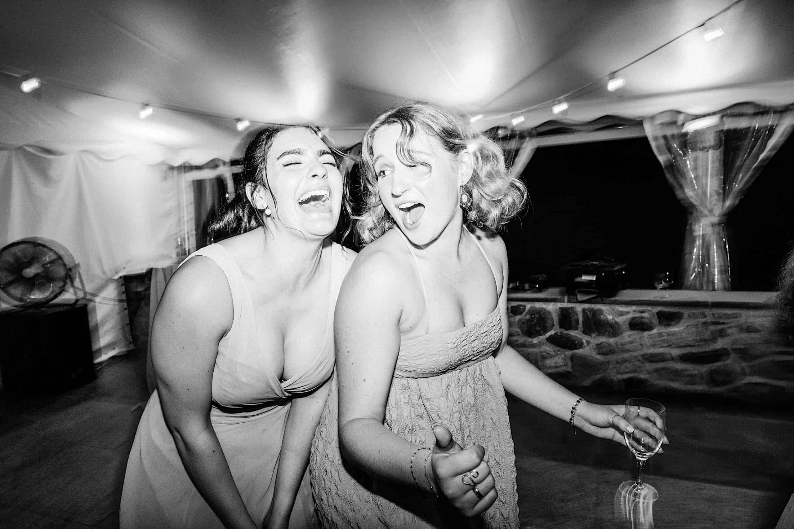 Two women in dresses laugh and pose for the camera at a Summer Wedding at Appleford Estate, one holding a wine glass. The festive scene features string lights and a charming stone wall in the background.