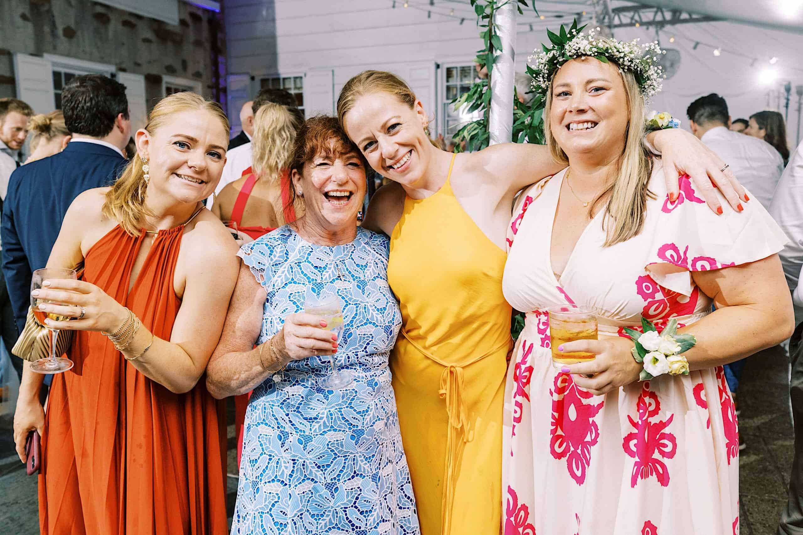 Four women smiling and posing together at a Summer Wedding at Appleford Estate, each holding a drink; one wears a floral crown and another dazzles in a yellow dress, capturing the joy of the celebration.