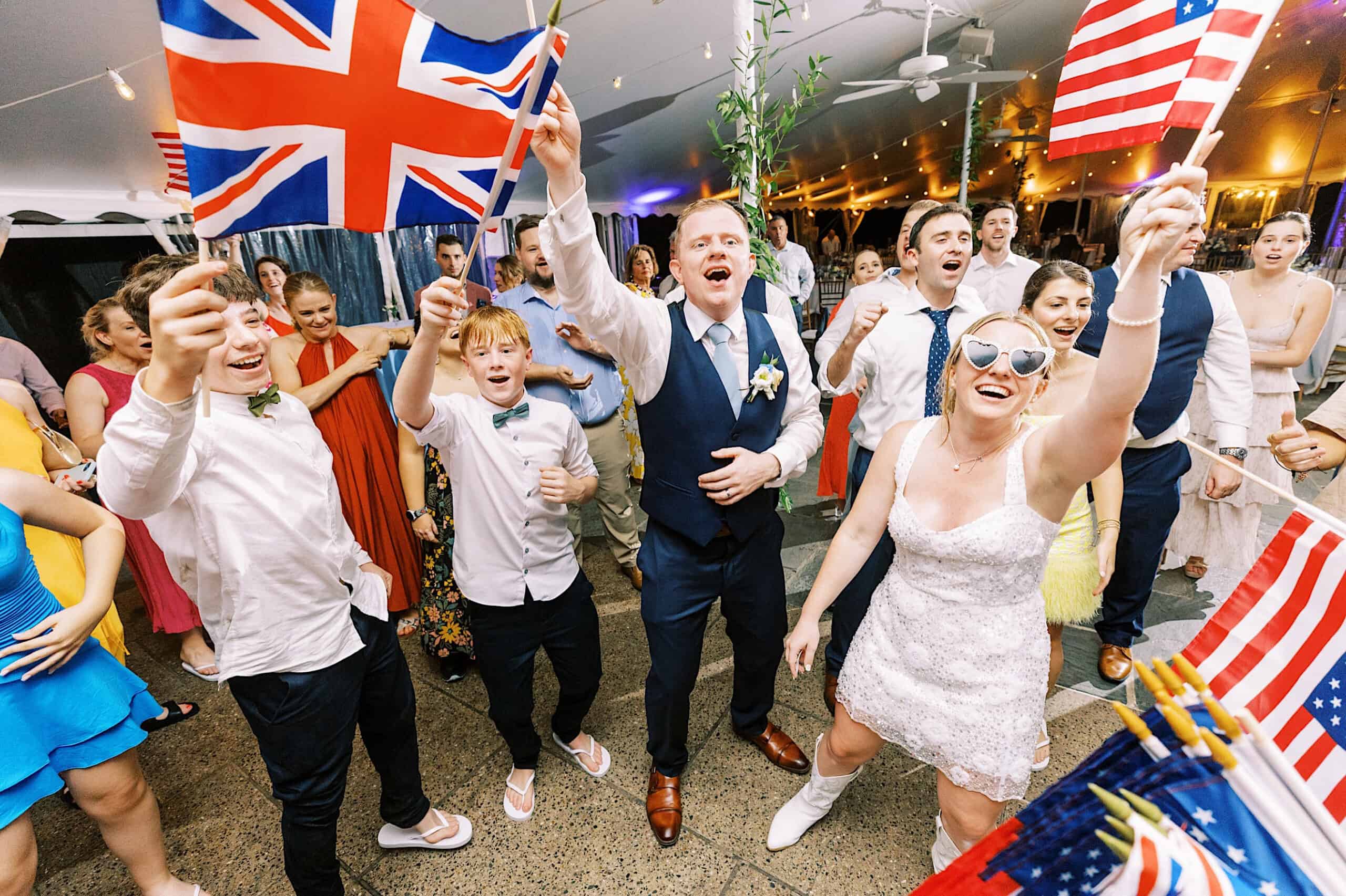 A group of people cheer and wave British and American flags at an indoor event reminiscent of a Summer Wedding at Appleford Estate; some are dressed formally, and one woman in white sunglasses holds a small flag.