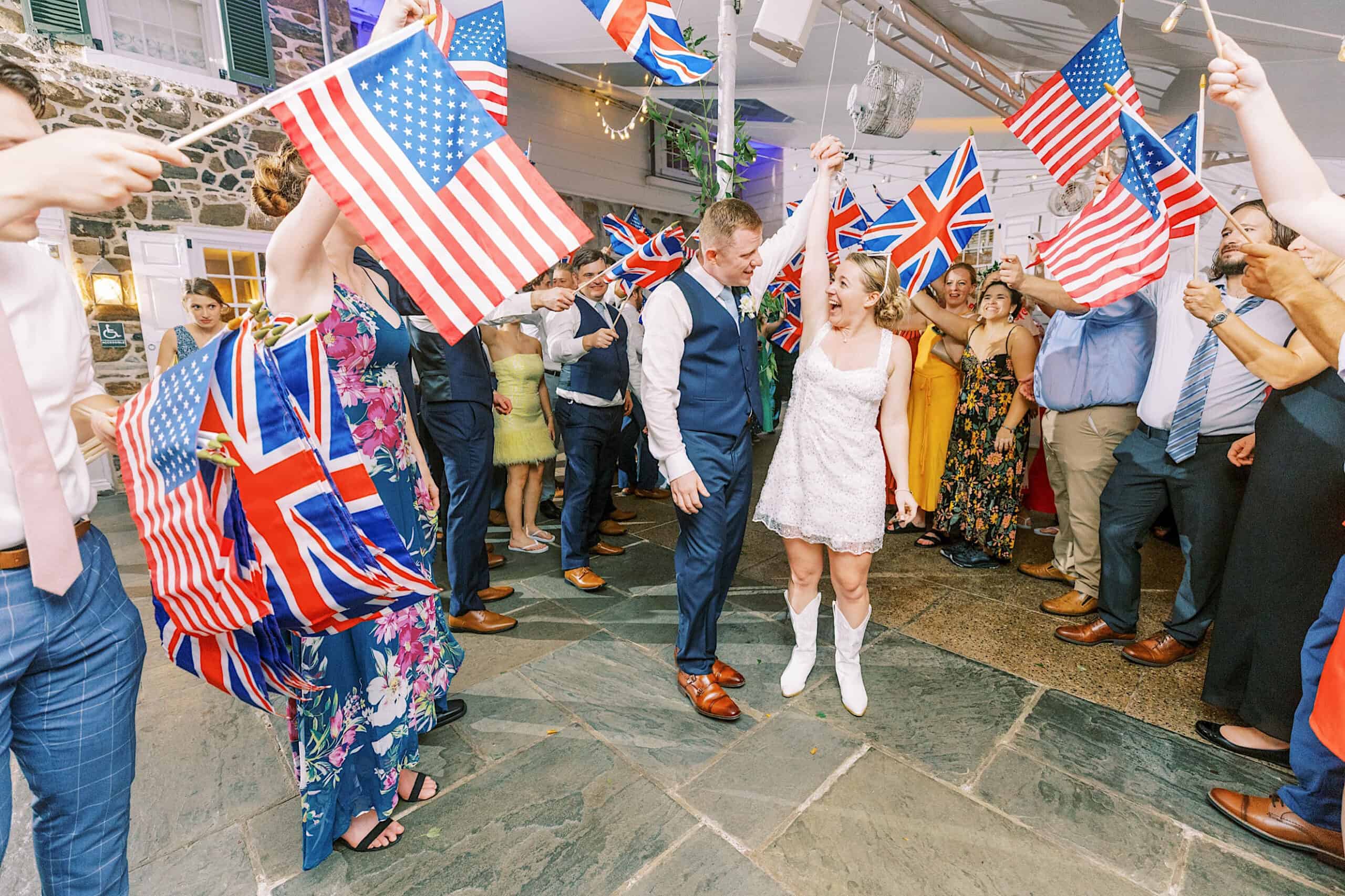 A couple walks through a crowd waving American and British flags at an indoor event, reminiscent of a Summer Wedding at Appleford Estate, with onlookers dressed in formal and semi-formal attire.