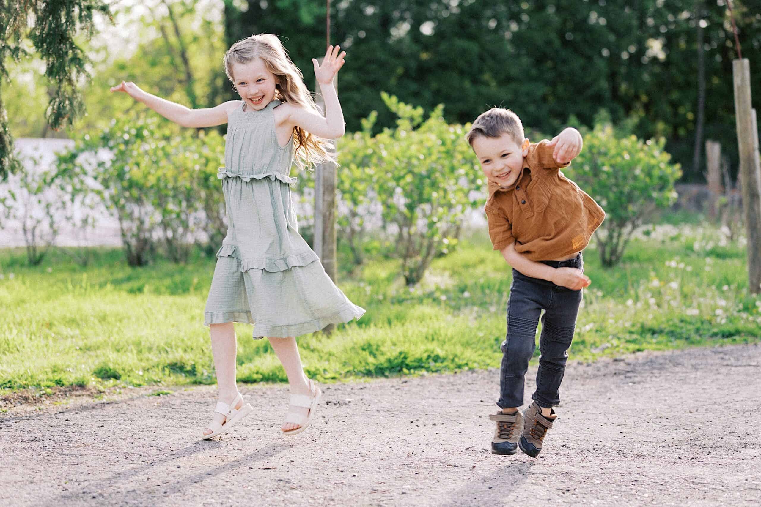 Two young children, a girl in a light dress and a boy in a brown shirt, are jumping and smiling outdoors on a sunny day with greenery in the background.