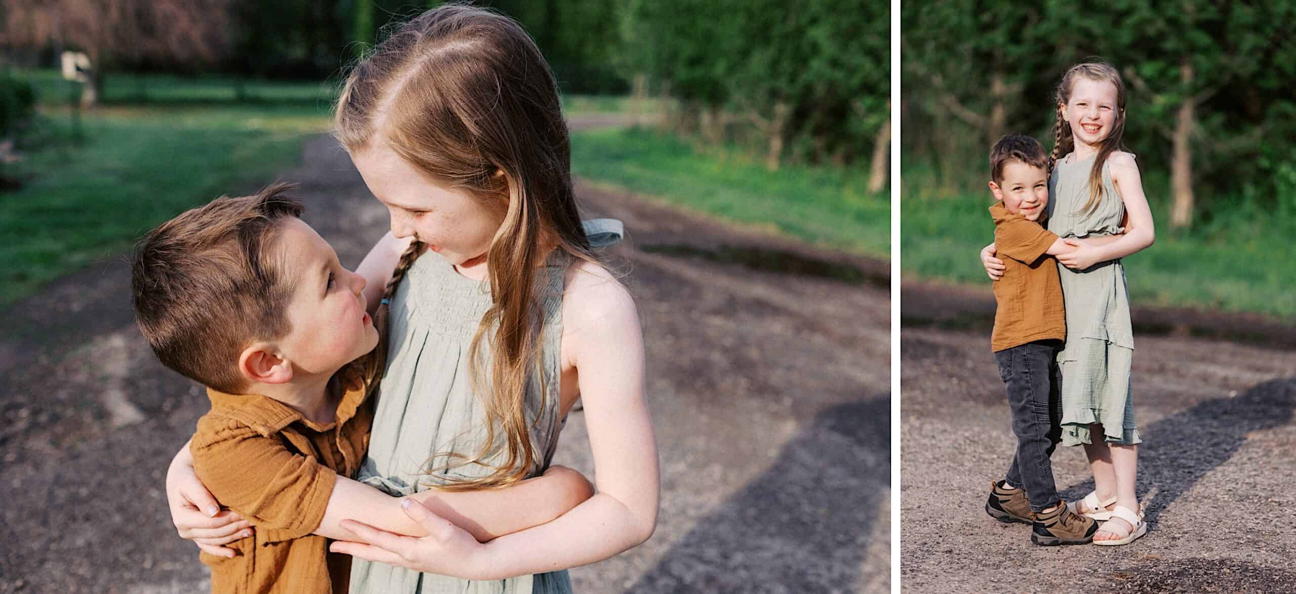 A young girl in a light green dress hugs a younger boy in a brown shirt while standing on a sunlit outdoor path surrounded by greenery.
