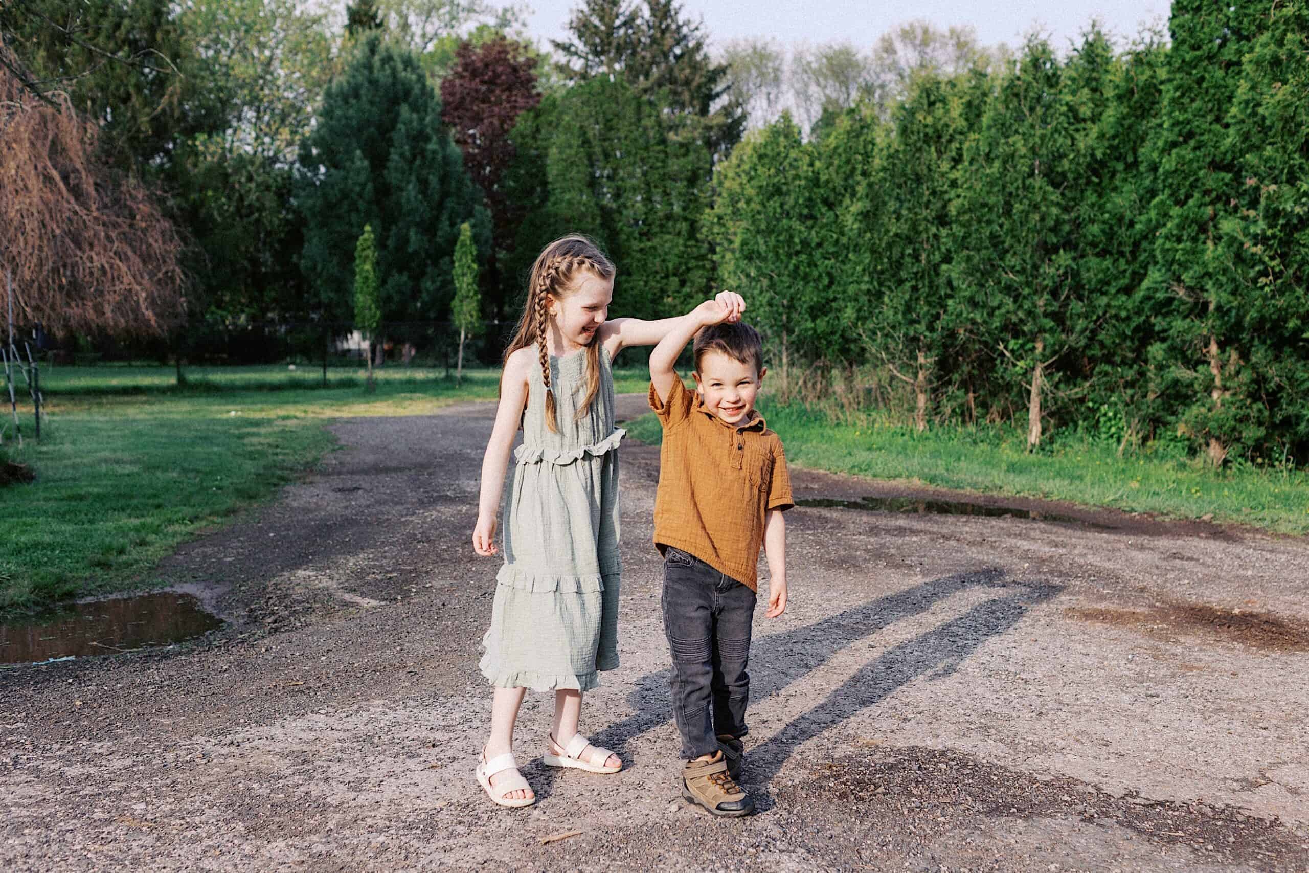 A girl in a light green dress holds up the hand of a smiling boy in a brown shirt as they stand on a gravel path in a park surrounded by trees.