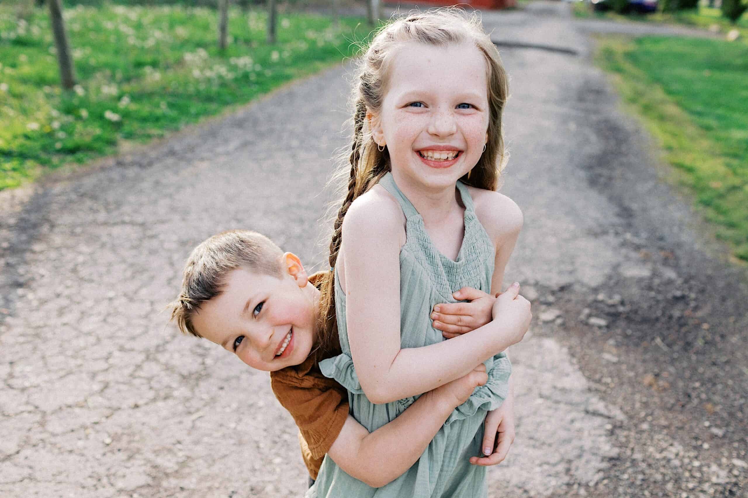 Two young children stand on a paved path outdoors; the boy hugs the girl from behind while both smile at the camera.