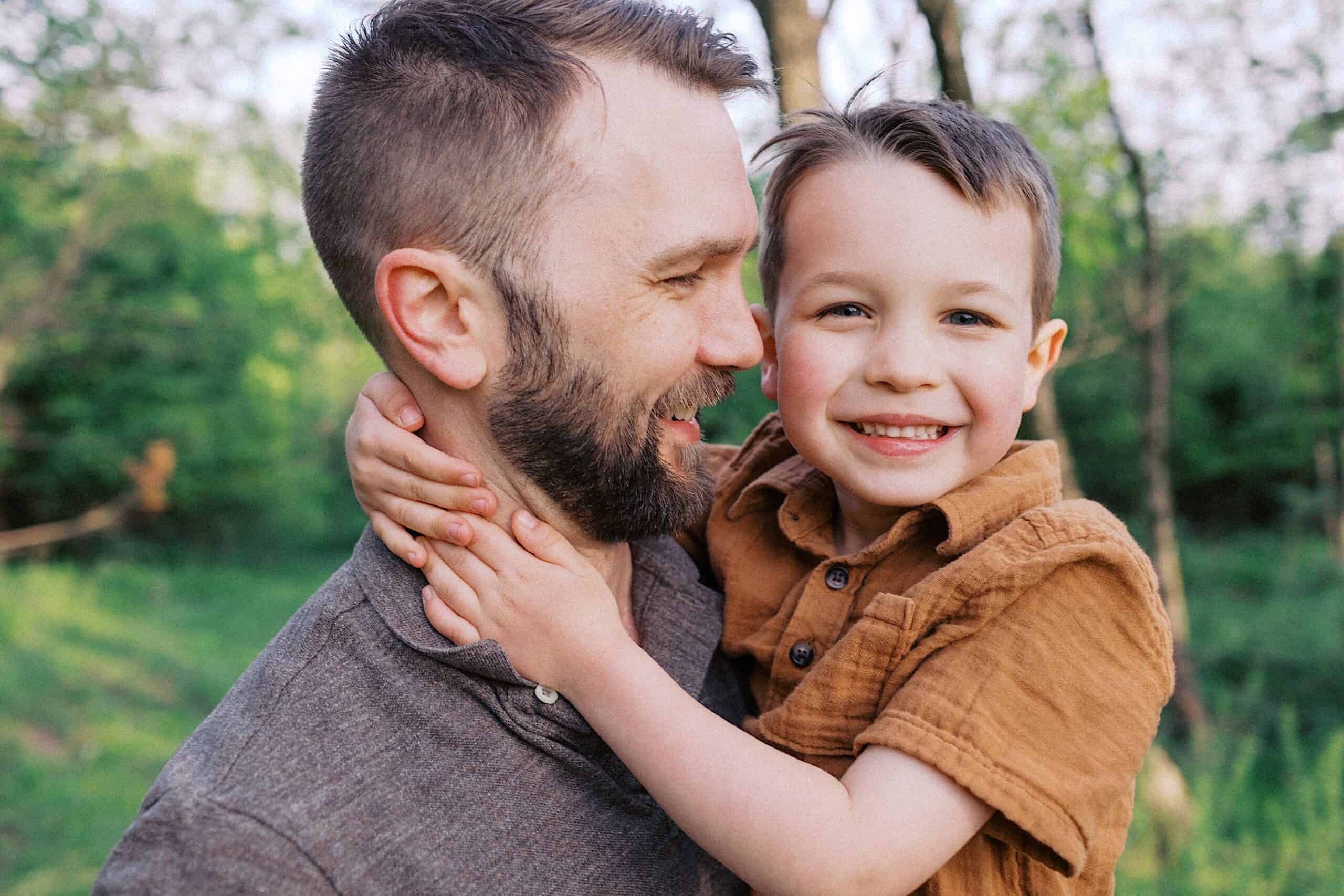 An adult man holds a smiling young boy outdoors in a green, wooded area. The boy has his arms around the man's neck, and both appear happy.