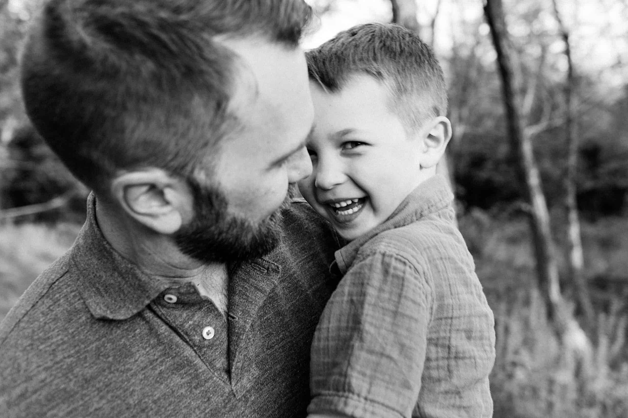 A man holds a smiling young boy outdoors, both wearing button-up shirts. The background is blurred with trees and grass.