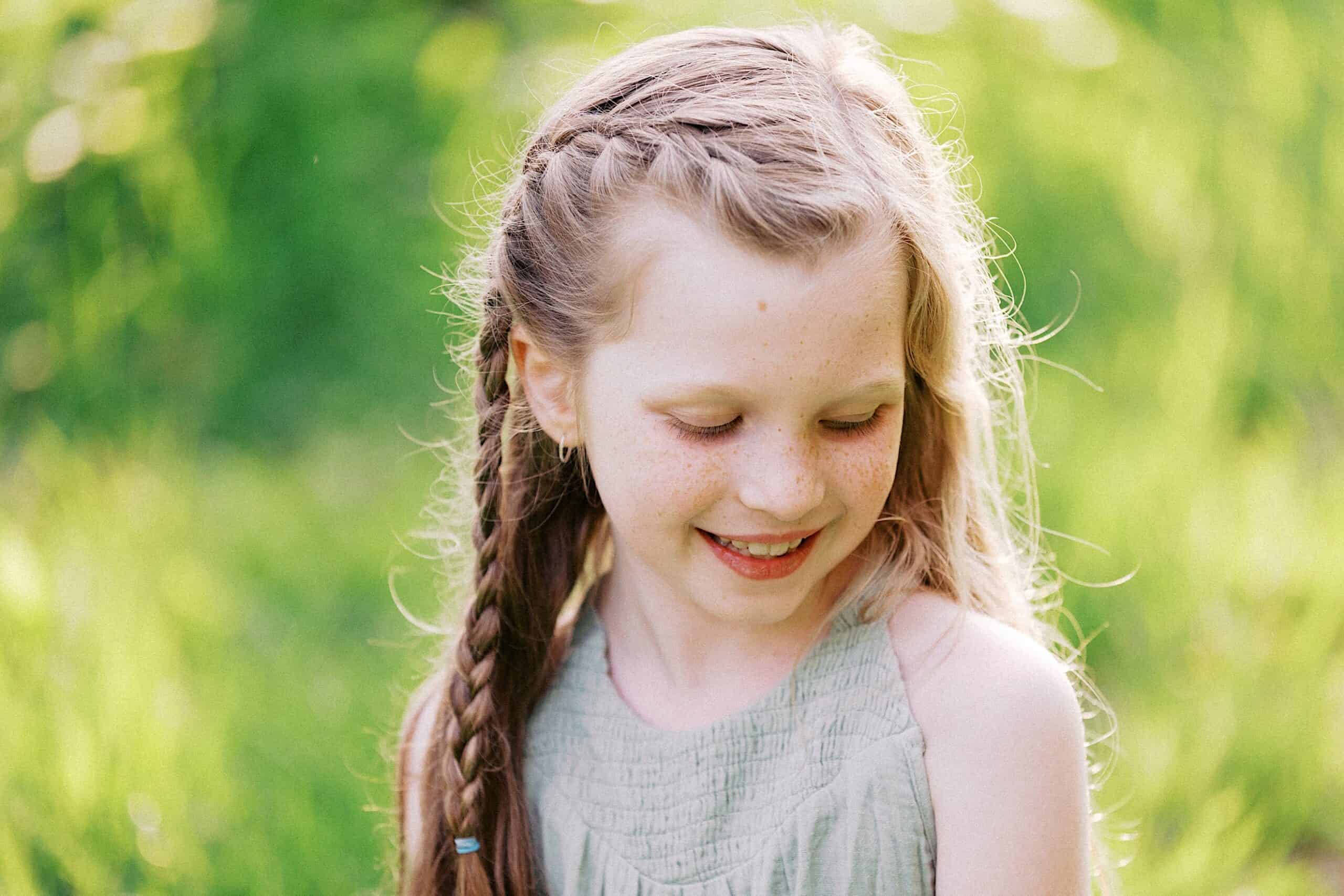 A young girl with long, braided hair and freckles smiles while looking down, standing outdoors in a sunlit grassy area.