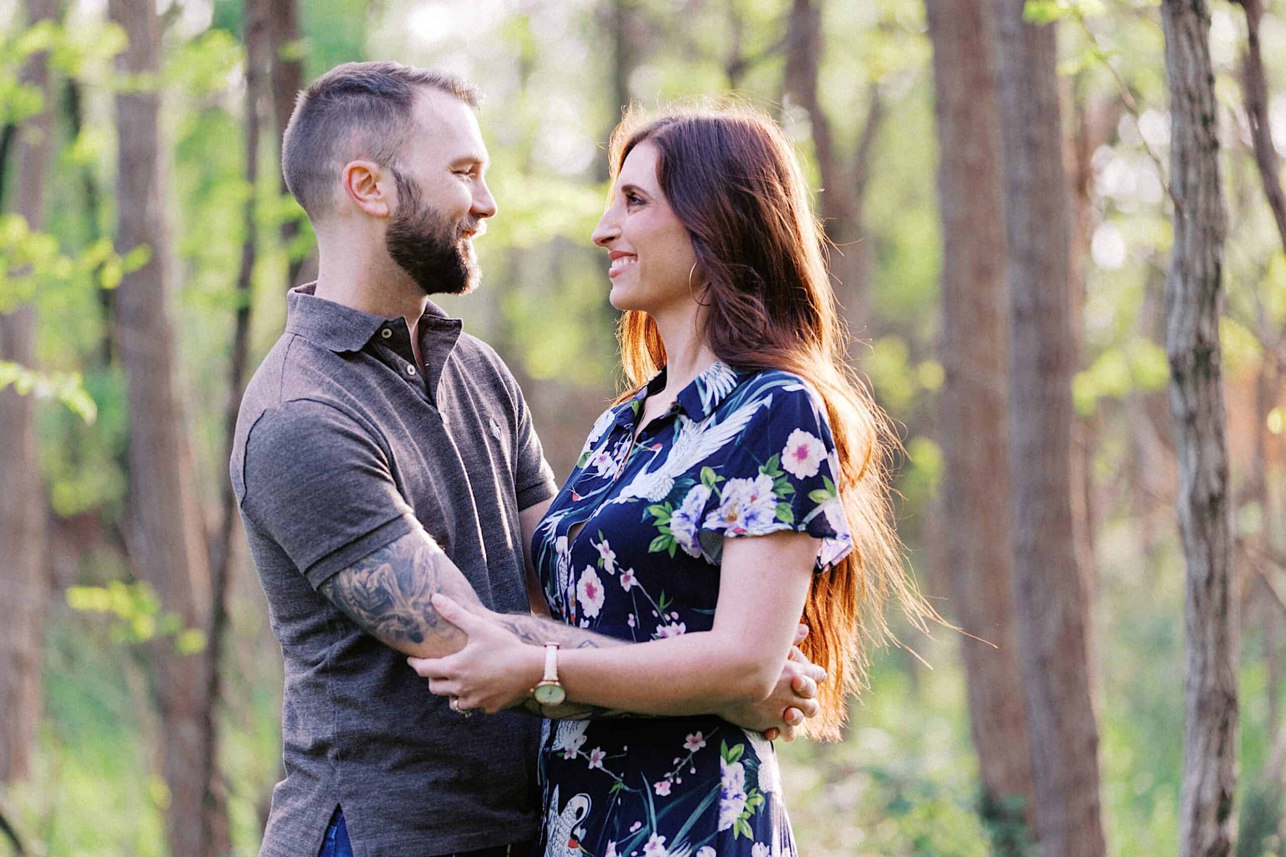 A man and woman stand in a forest, facing each other and smiling, with their arms around each other. The background is filled with trees and greenery.