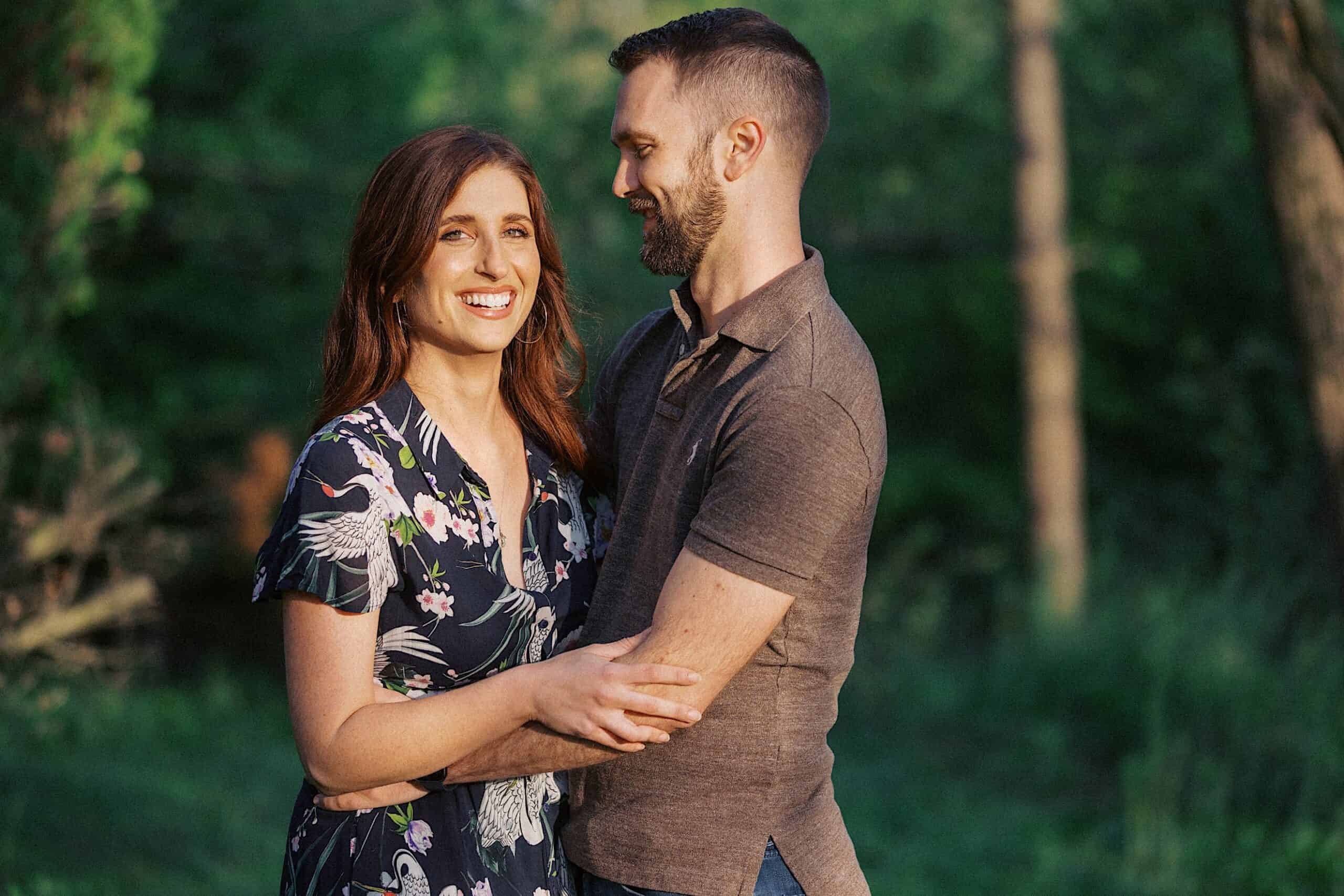 A woman in a floral dress and a man in a brown shirt stand close together outdoors, smiling and holding each other, with greenery in the background.