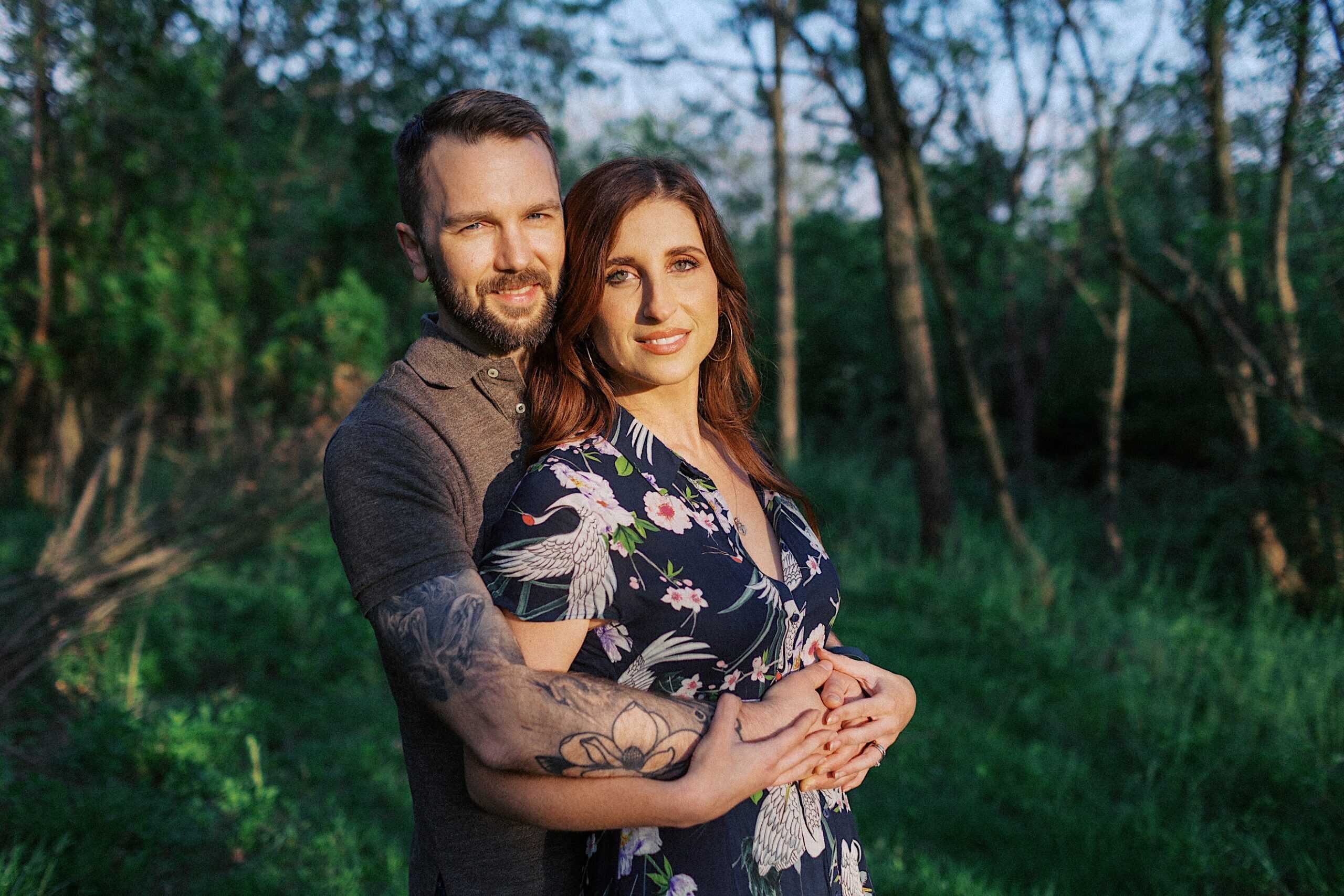 A man with tattoos hugs a woman from behind in a sunlit forest clearing. Both are looking at the camera and smiling. The woman wears a floral dress.