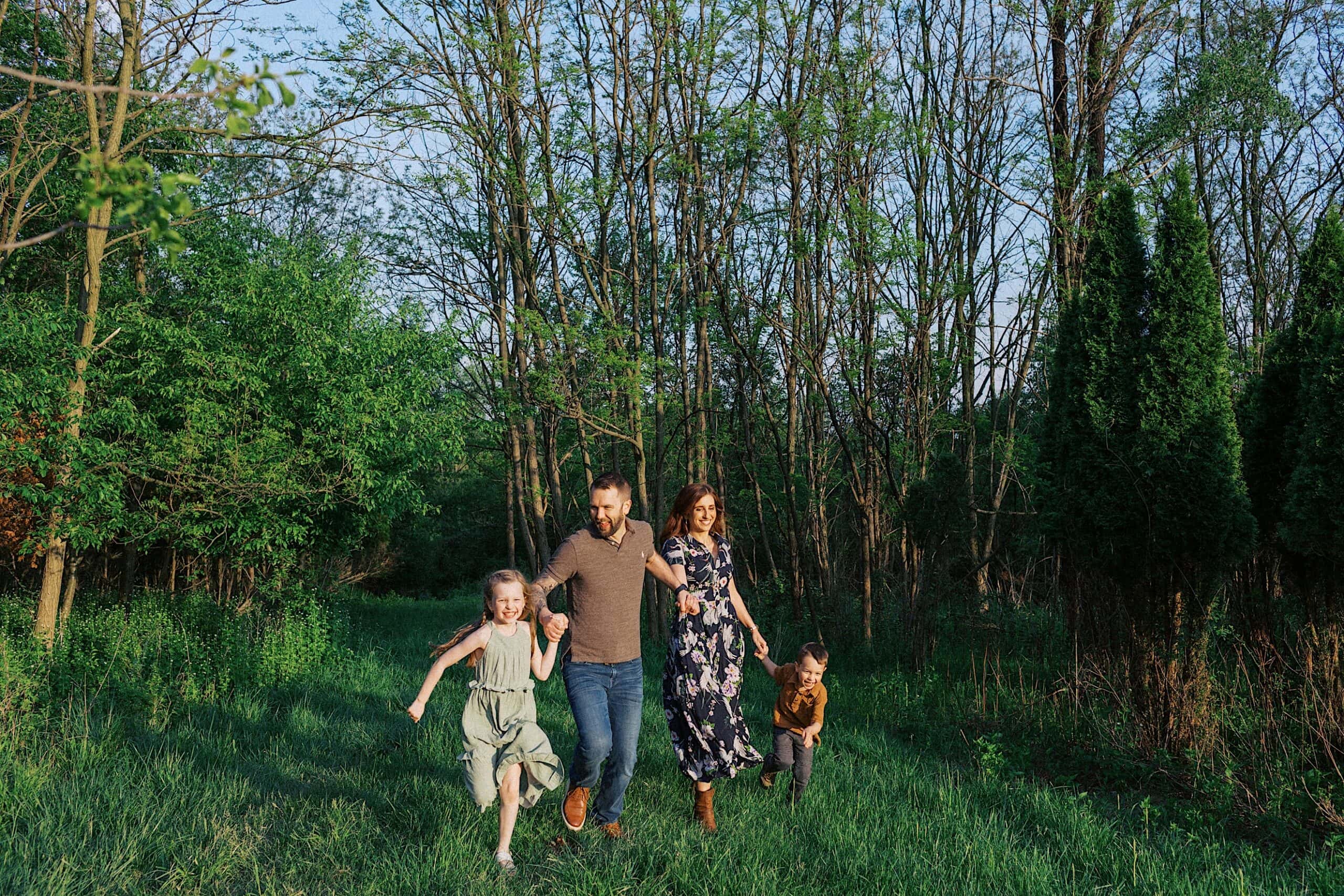 A family of four—two adults and two children—holds hands and runs through a grassy area surrounded by trees.