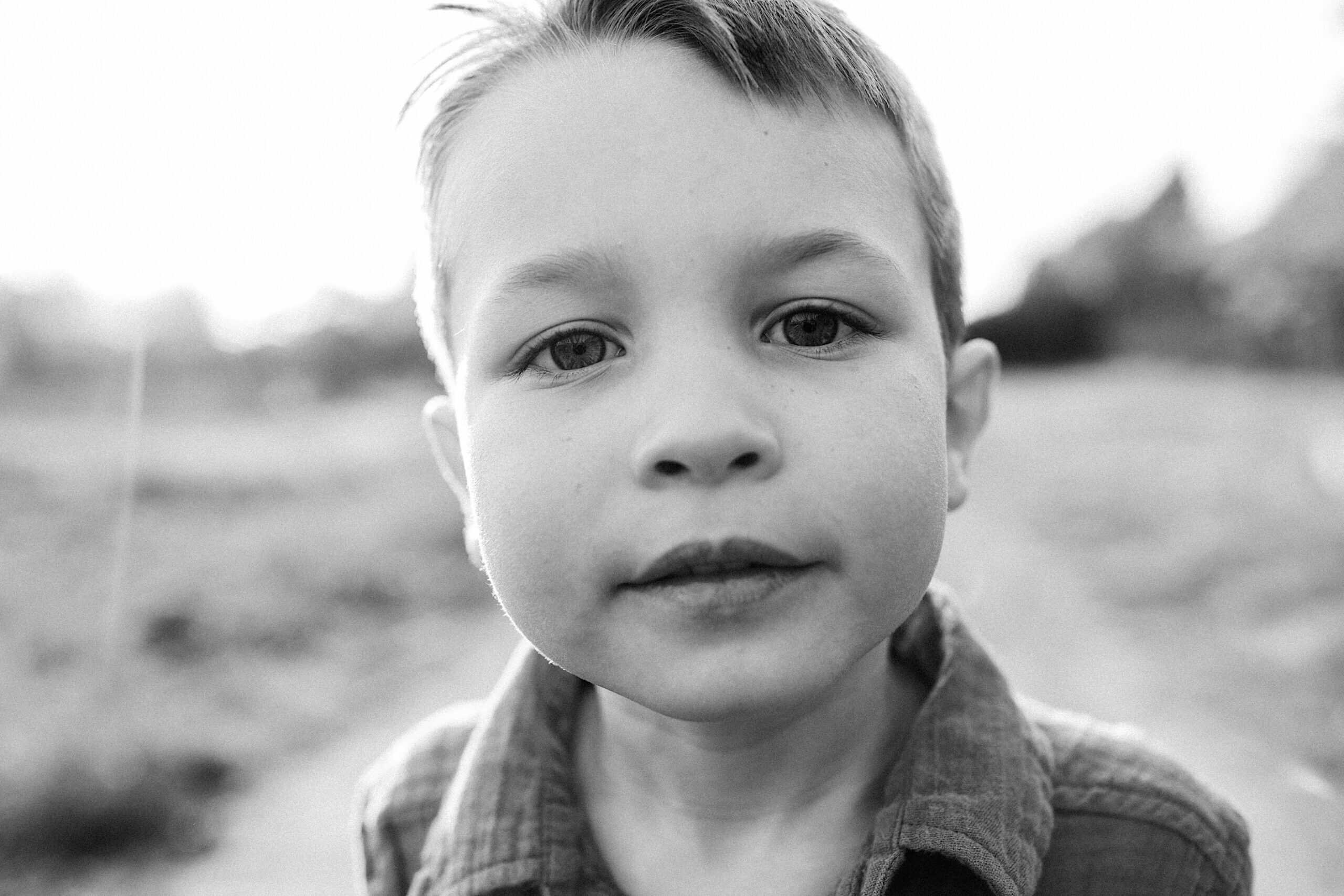 Black and white close-up of a young boy outdoors, looking directly at the camera, with a blurred background.