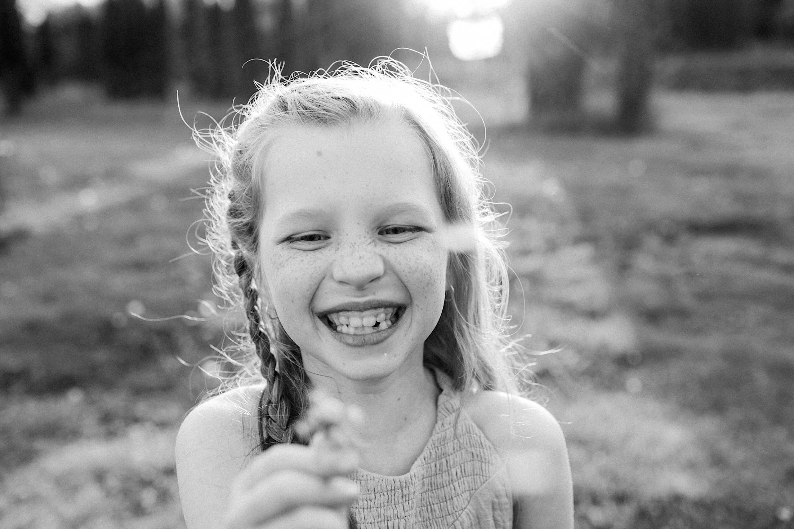 A young girl with braids and freckles smiles outdoors, holding a small object up to the camera. The image is in black and white with sunlight in the background.
