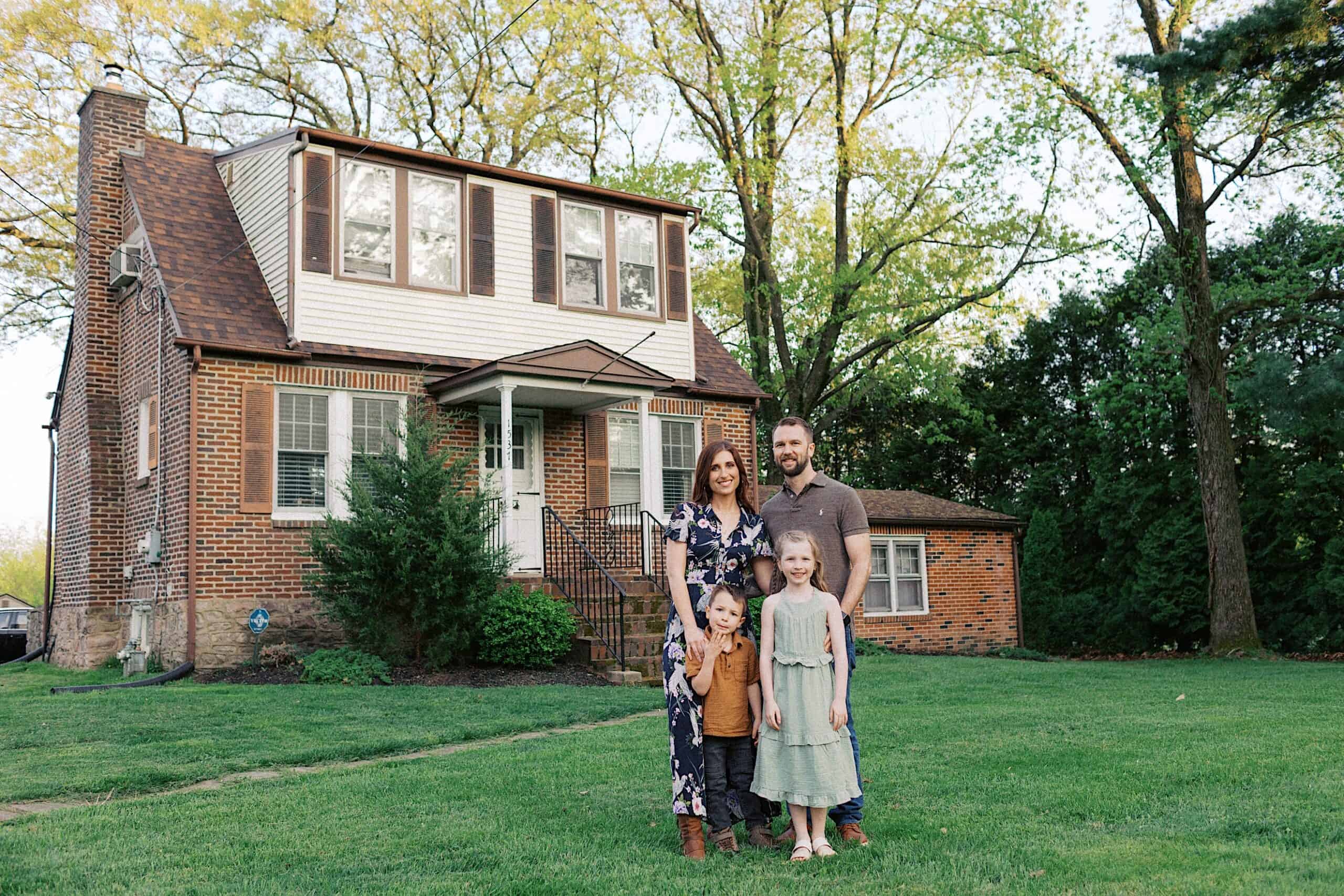 A family of four stands together on the lawn in front of a two-story brick house with trees and shrubbery in the background.