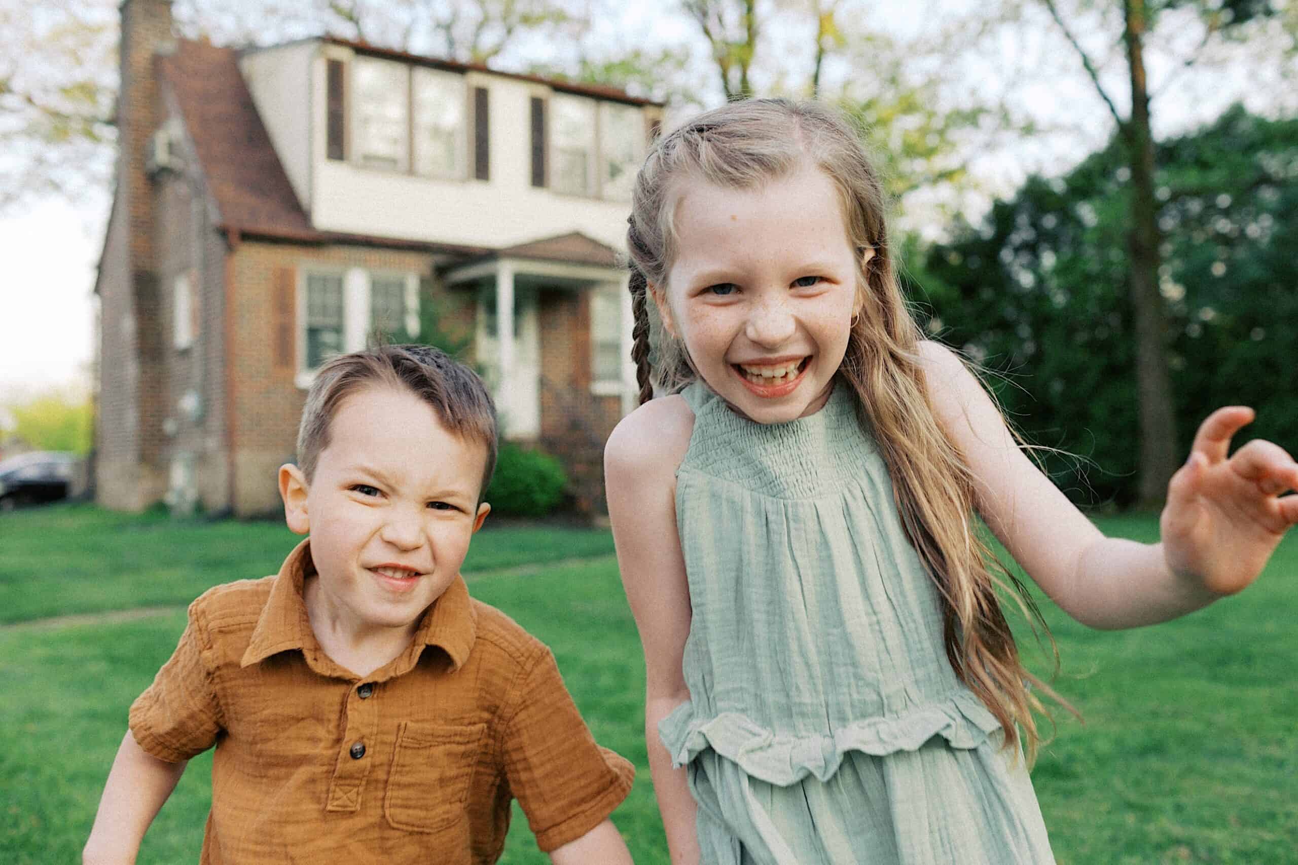 A young boy in a brown shirt and a young girl in a light dress are smiling and playing outside on a lawn in front of a brick house.