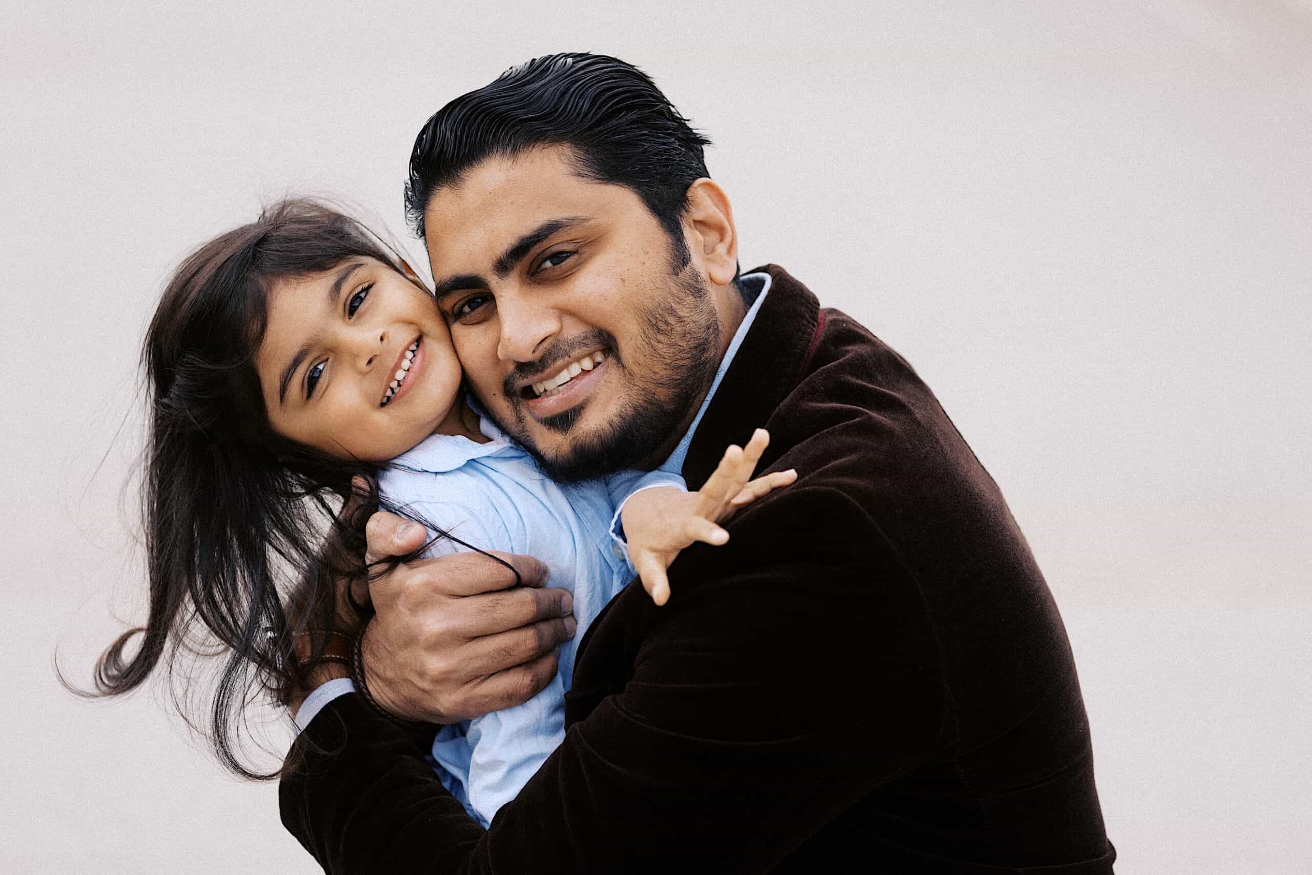 A man wearing a dark jacket hugs a smiling young girl with long hair. Both are looking at the camera against a plain background.