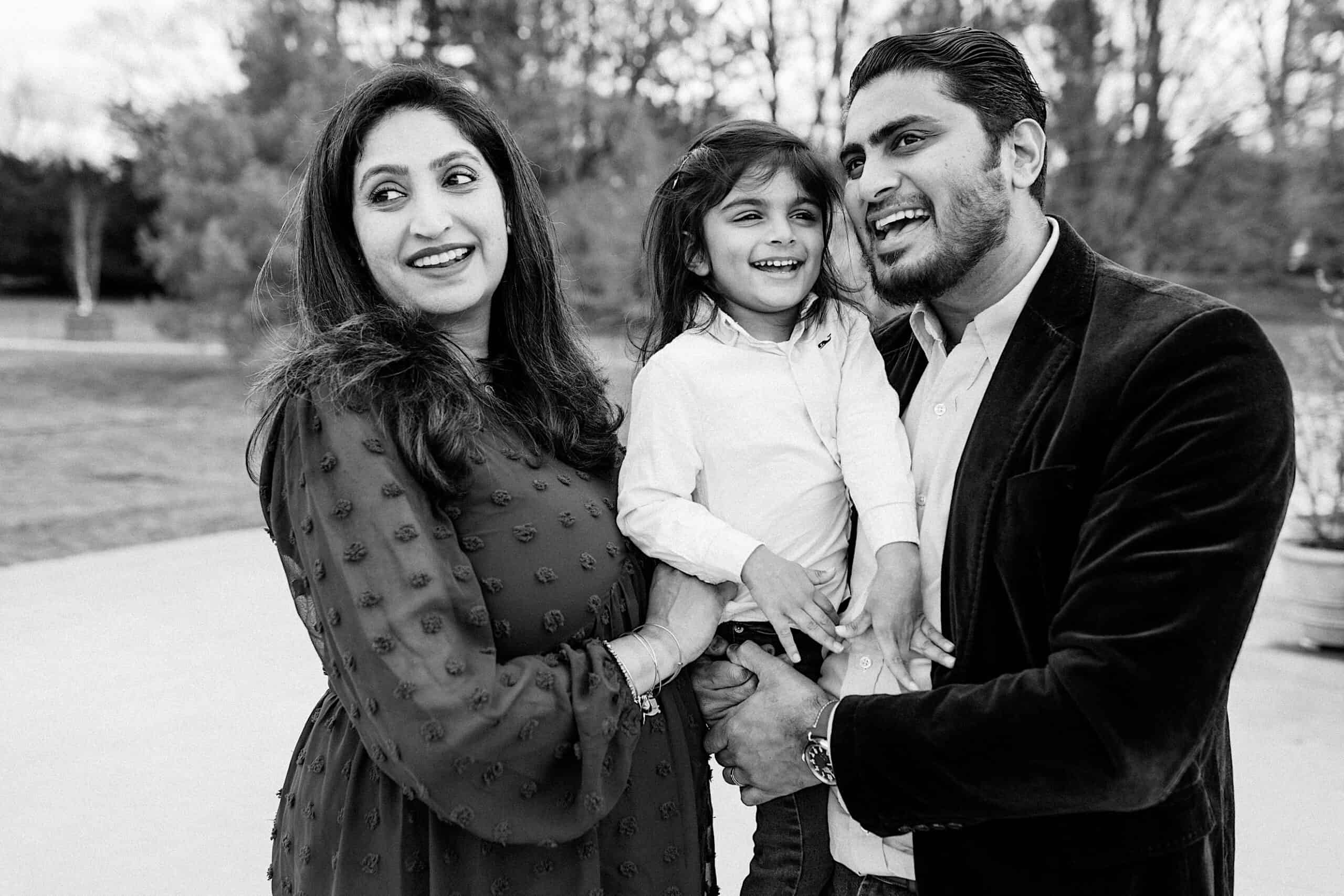 A man and woman stand outside holding a young child. All three are smiling and looking in different directions. Trees are visible in the background.