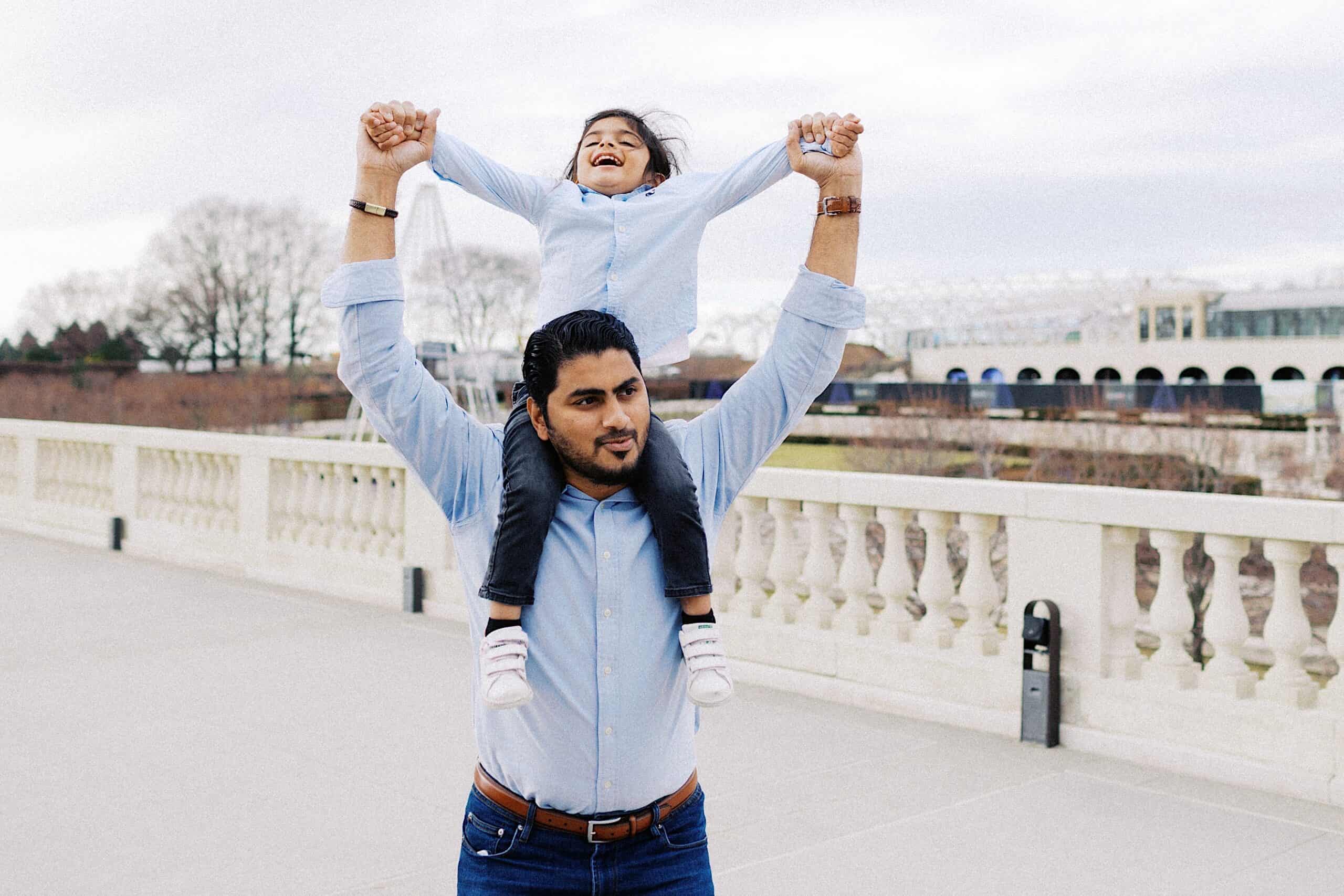 An adult carrying a smiling child on their shoulders while holding the child’s hands, outdoors on a paved walkway with a railing and trees in the background.