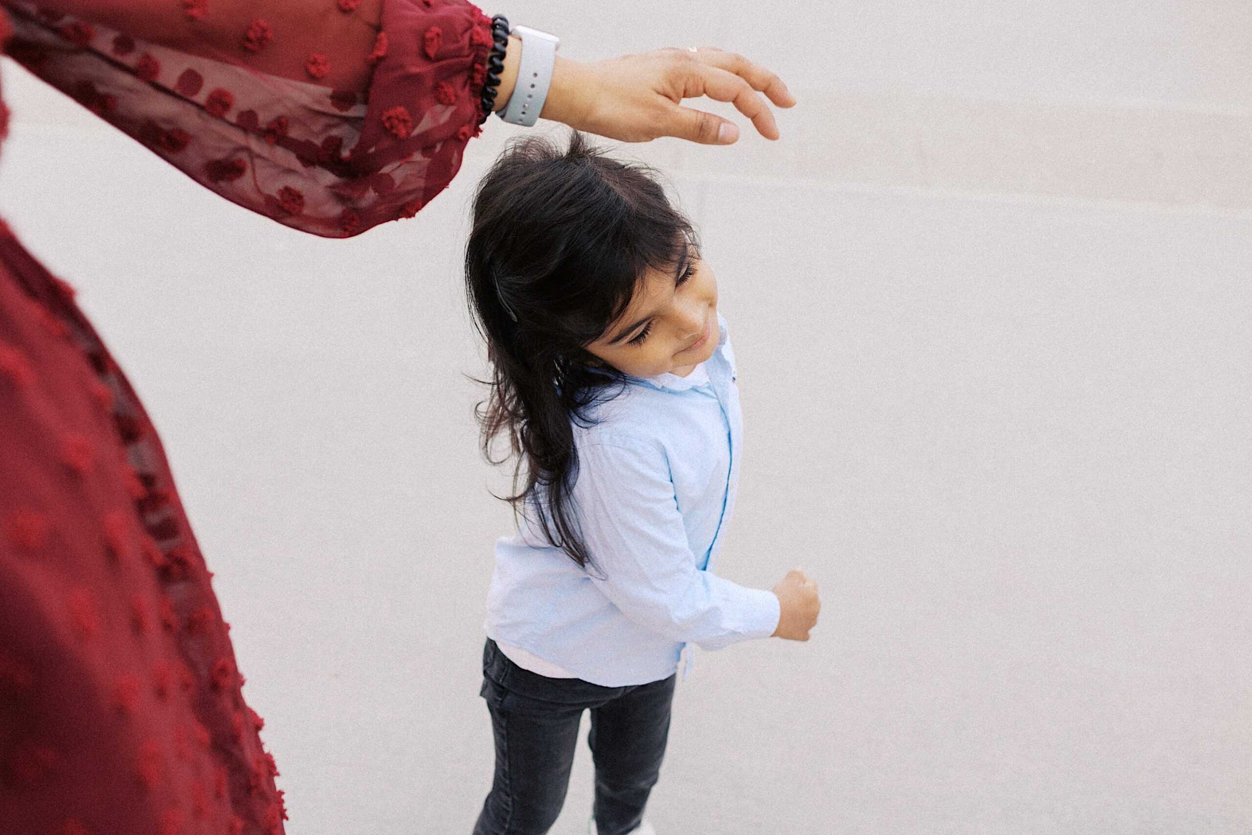 A young child in a light blue shirt and dark pants walks outdoors, looking up as an adult's hand reaches overhead.