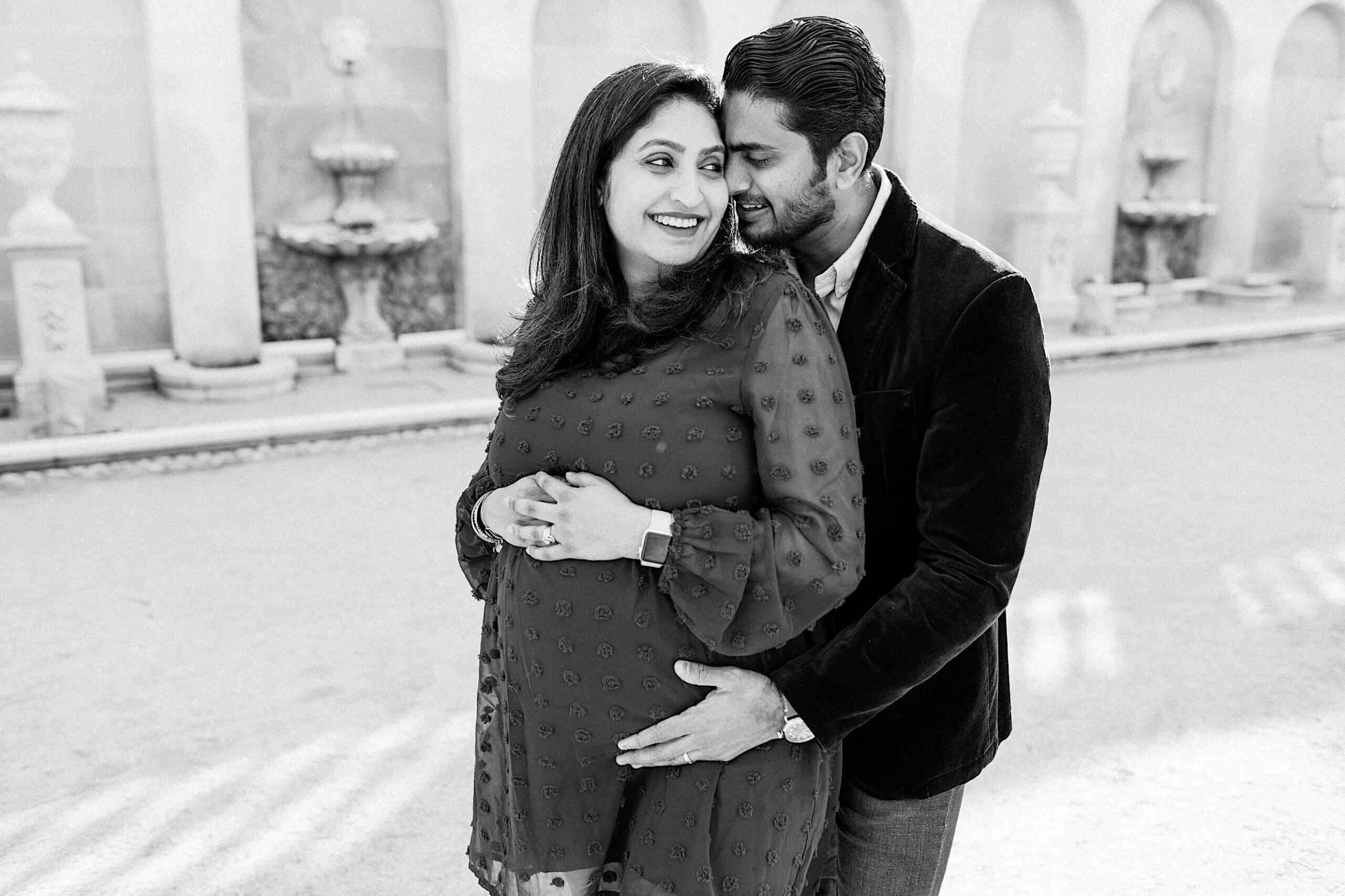 A man stands behind a pregnant woman, both smiling and holding her belly, in an outdoor area with stone fountains and arches in the background.