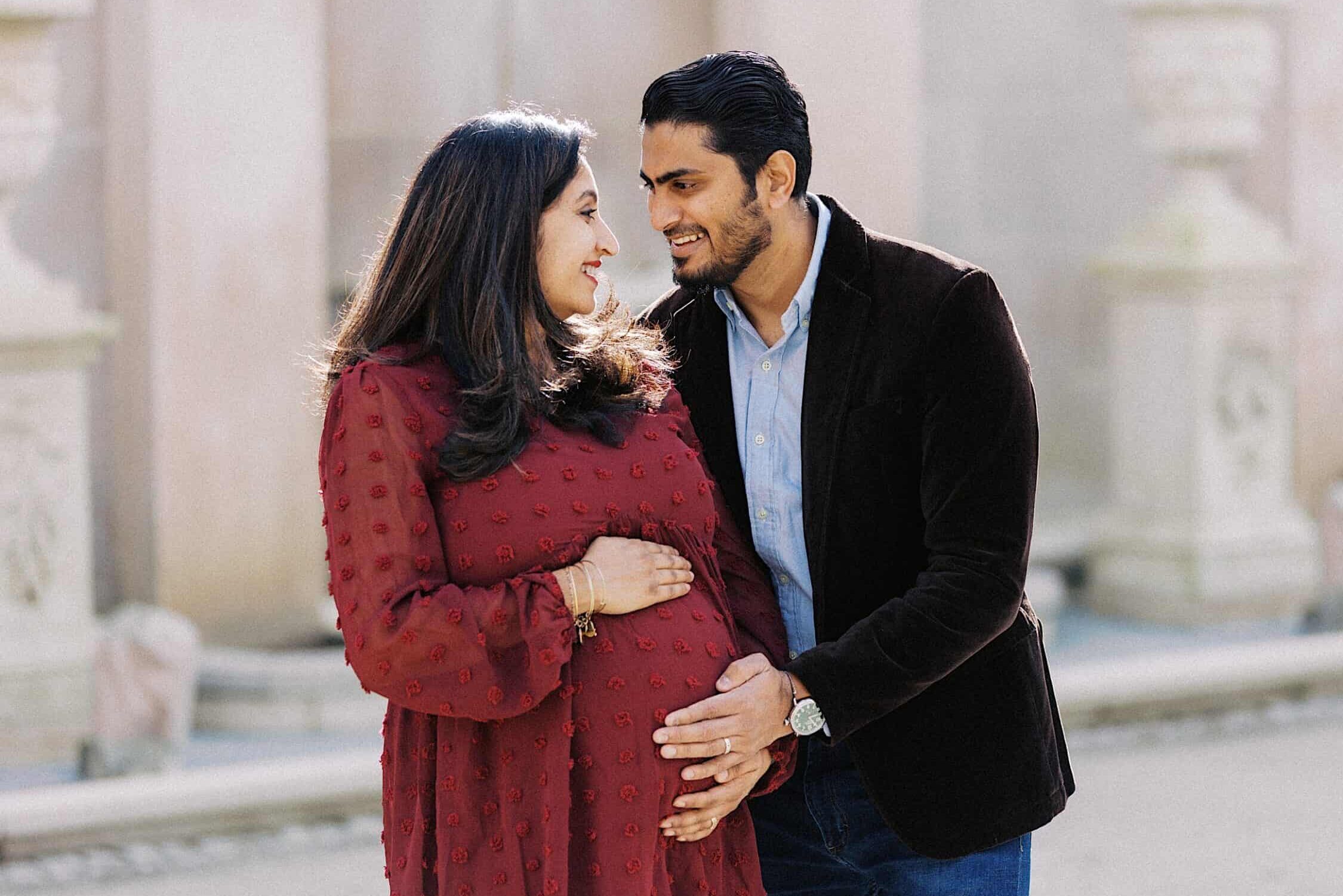A man and a pregnant woman stand close together outdoors, smiling at each other. The man gently places his hand on the woman's baby bump.