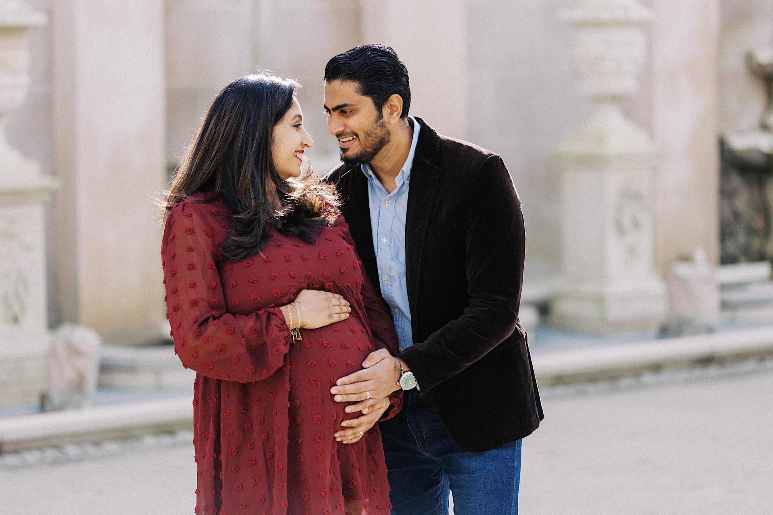 A man and a pregnant woman stand close together outdoors, smiling at each other. The man gently places his hand on the woman's baby bump.