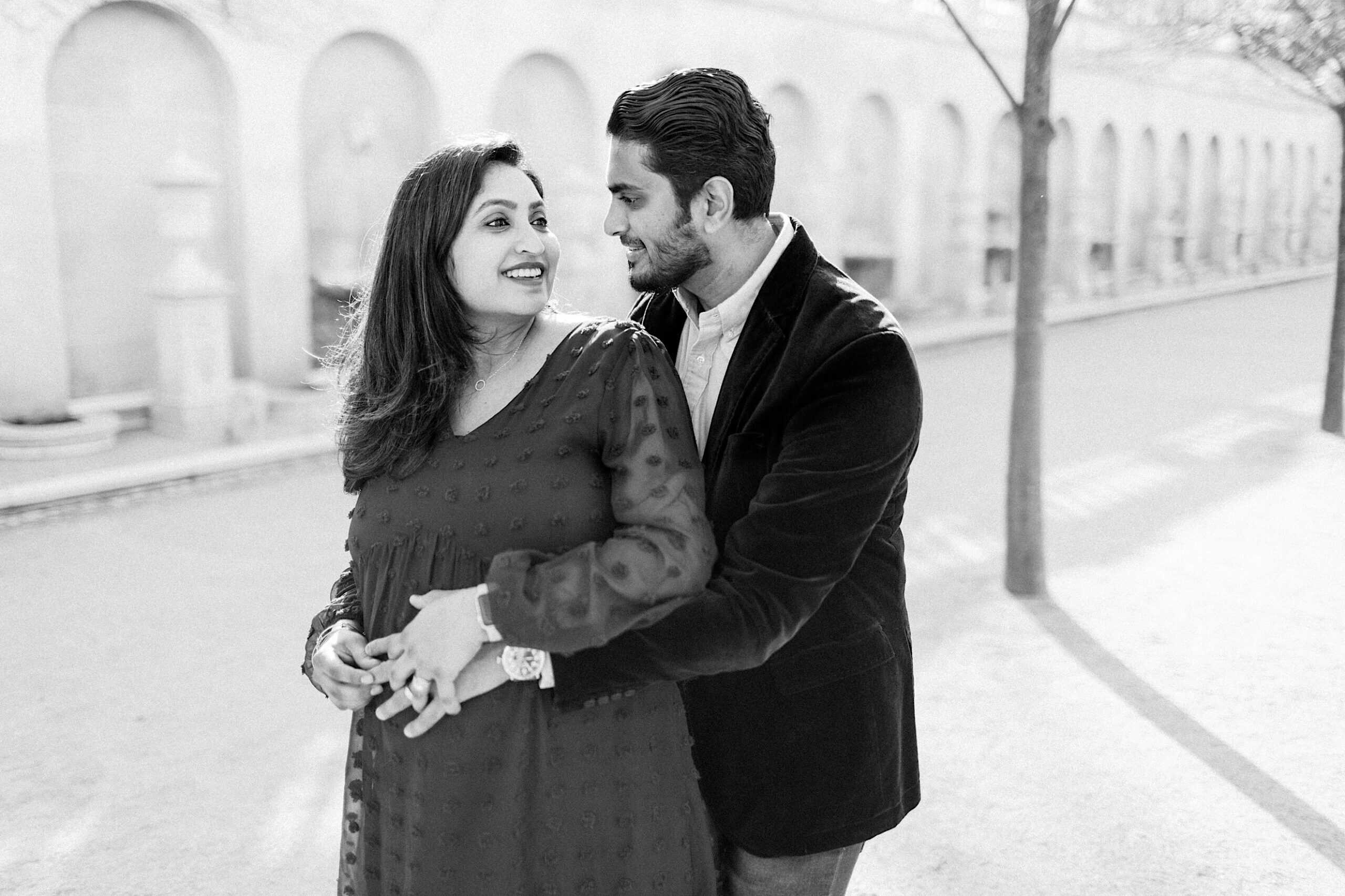 A man stands behind a woman, holding her around the waist as they smile at each other on a sunlit, tree-lined path.