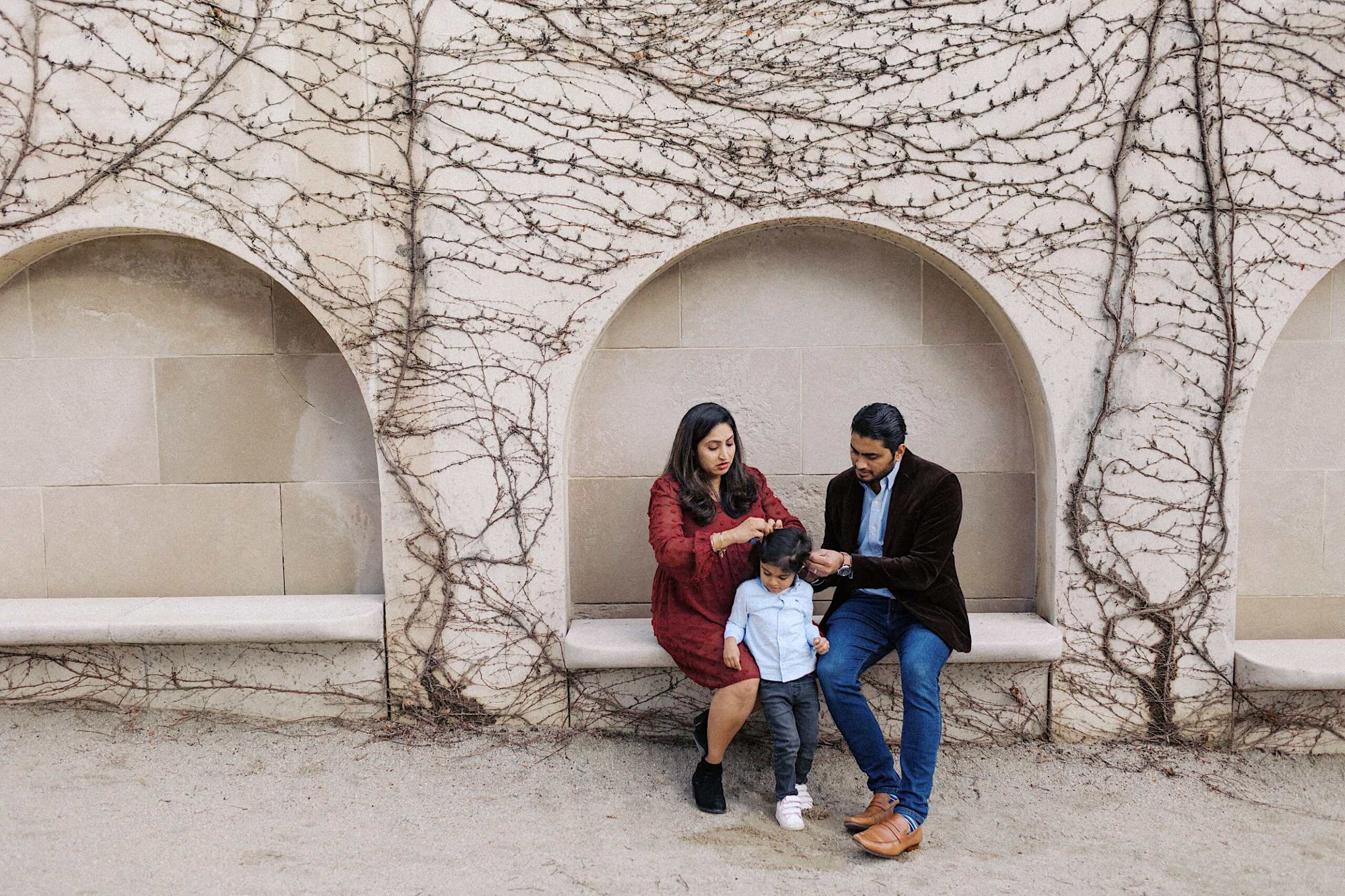 A woman and a man sit on a stone bench under an archway, helping a young child stand. Leafless vines climb the wall behind them.