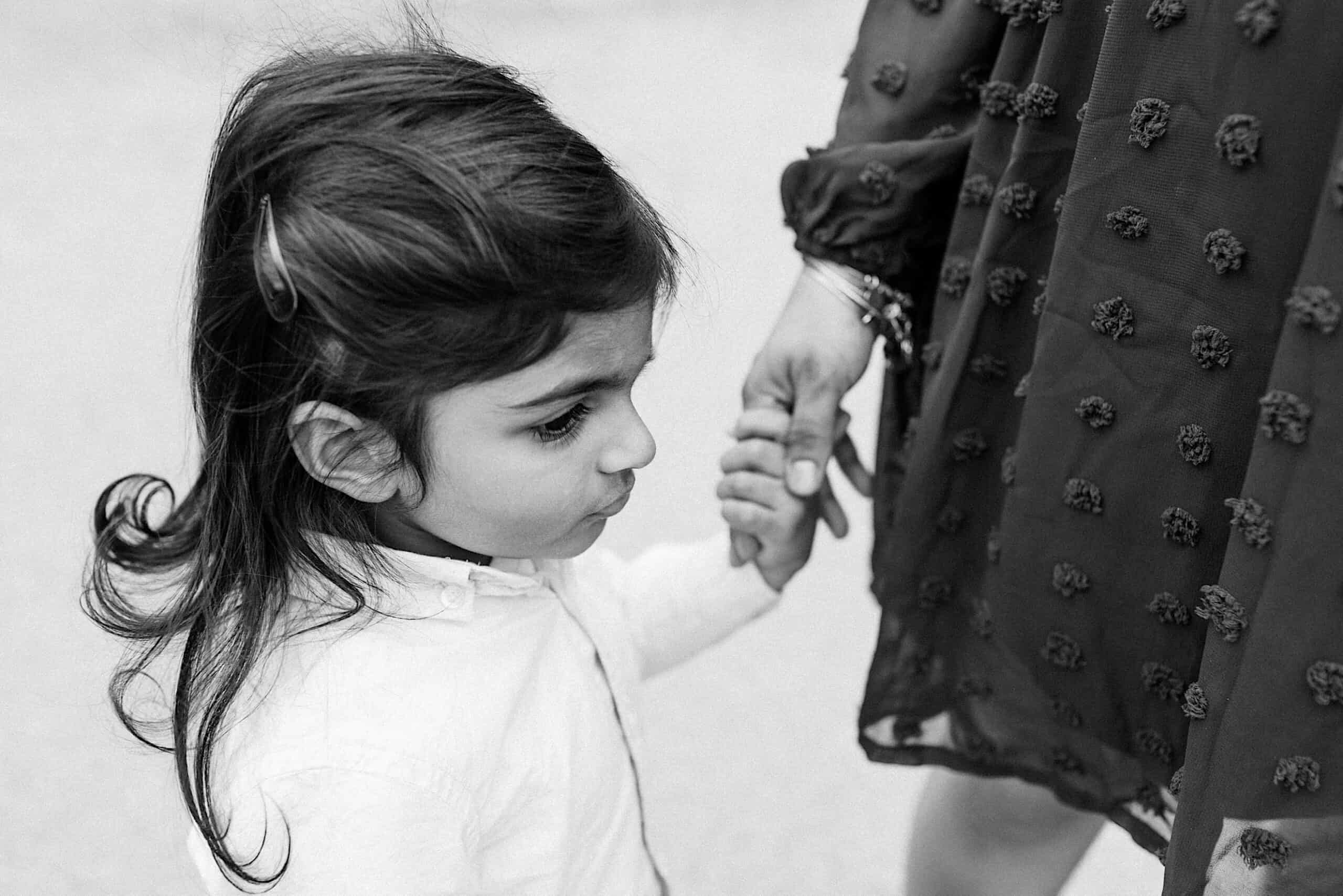 A young child with long hair holds an adult's hand, looking slightly to the side; the adult wears a textured dress.