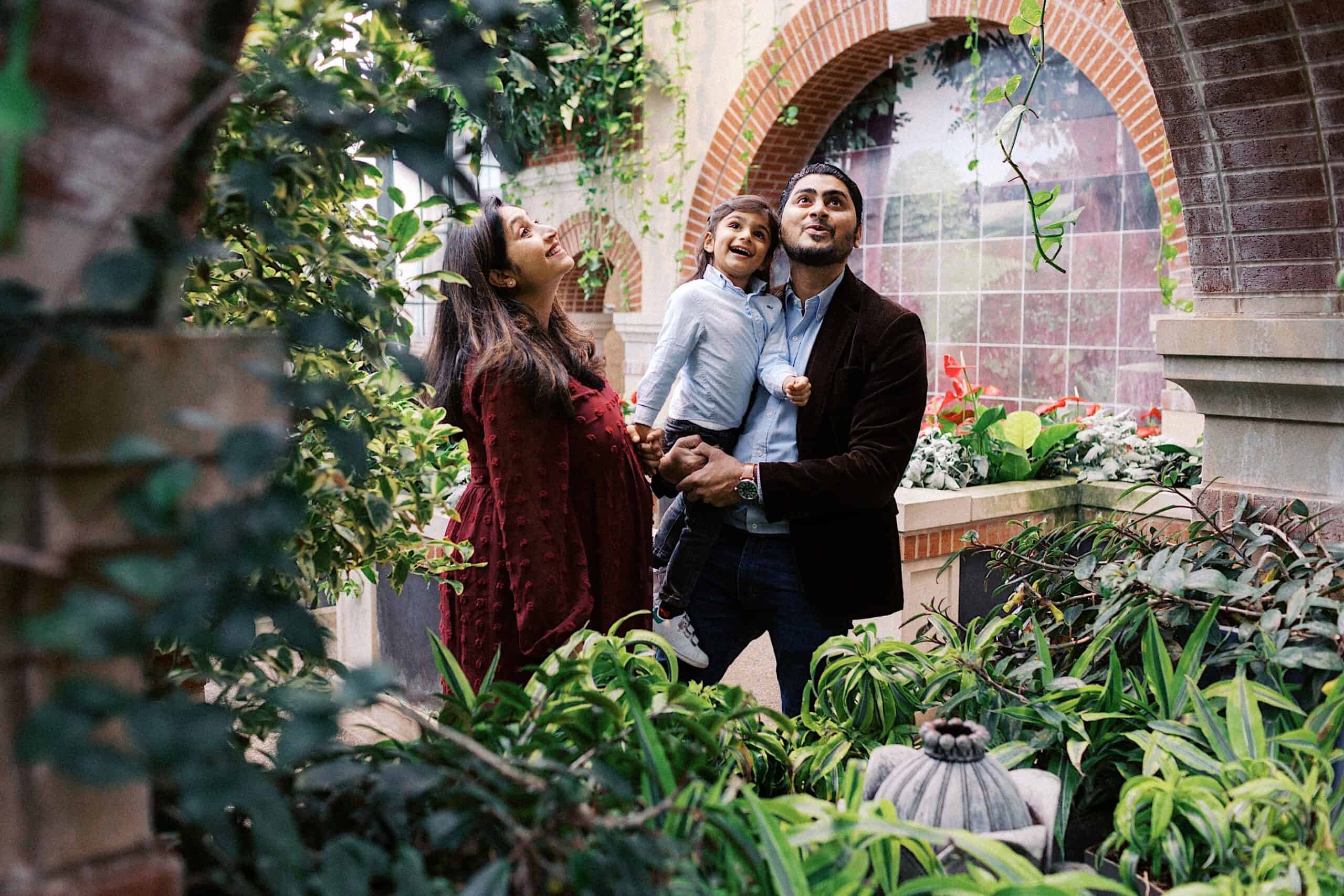 A family of three stands in a lush garden under brick arches; the parents hold their child, who is smiling and looking up.