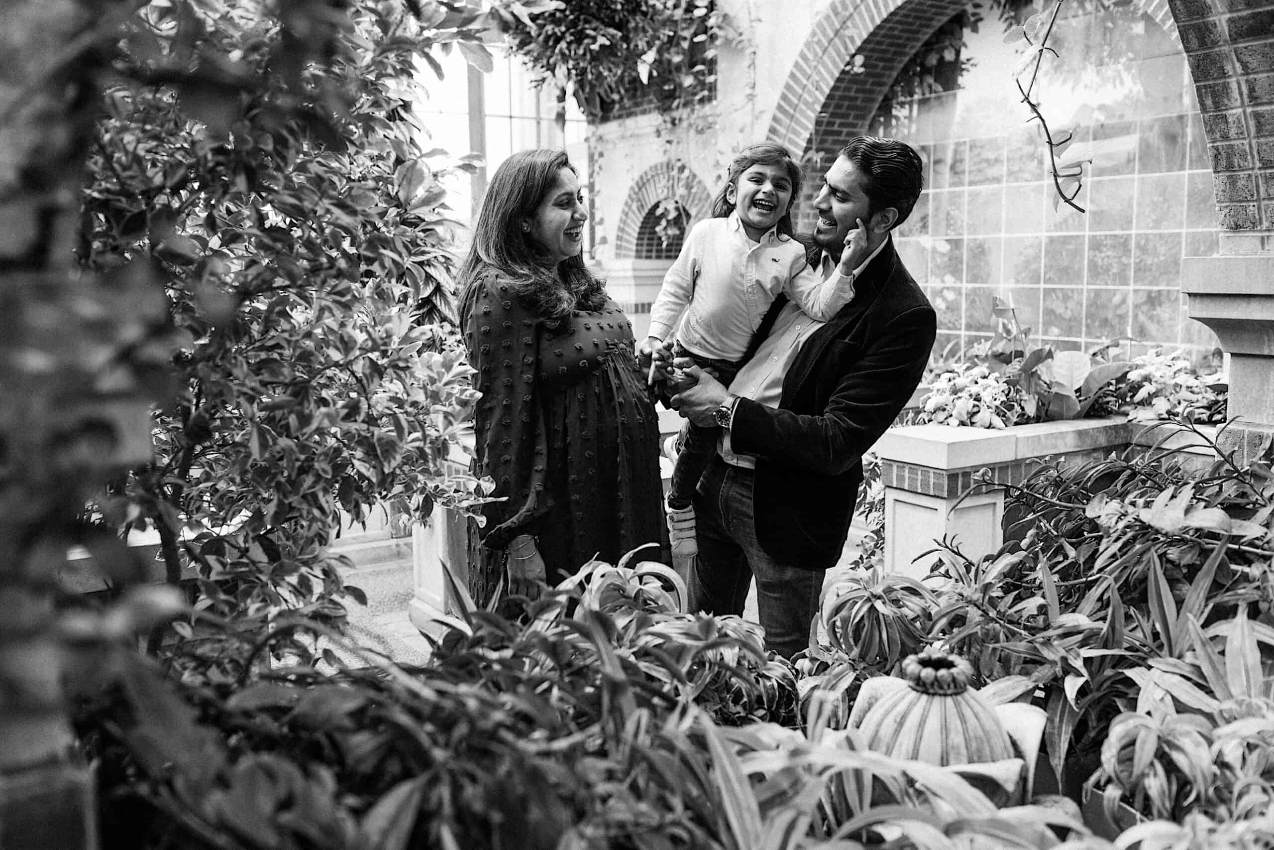 A man, woman, and child stand together, smiling among plants and greenery in an indoor garden setting.