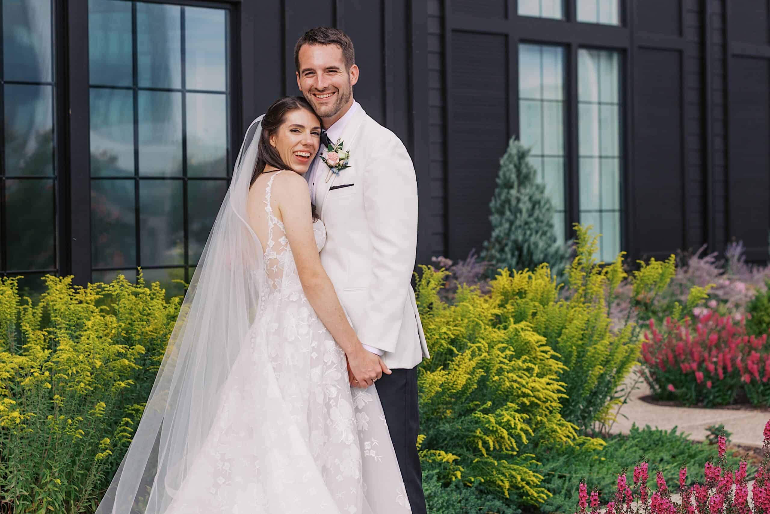 A bride and groom stand outdoors in front of a modern building with large windows, smiling and holding hands, surrounded by colorful flowers and greenery during their luxurious wedding at The Ivy at Ellis Preserve.